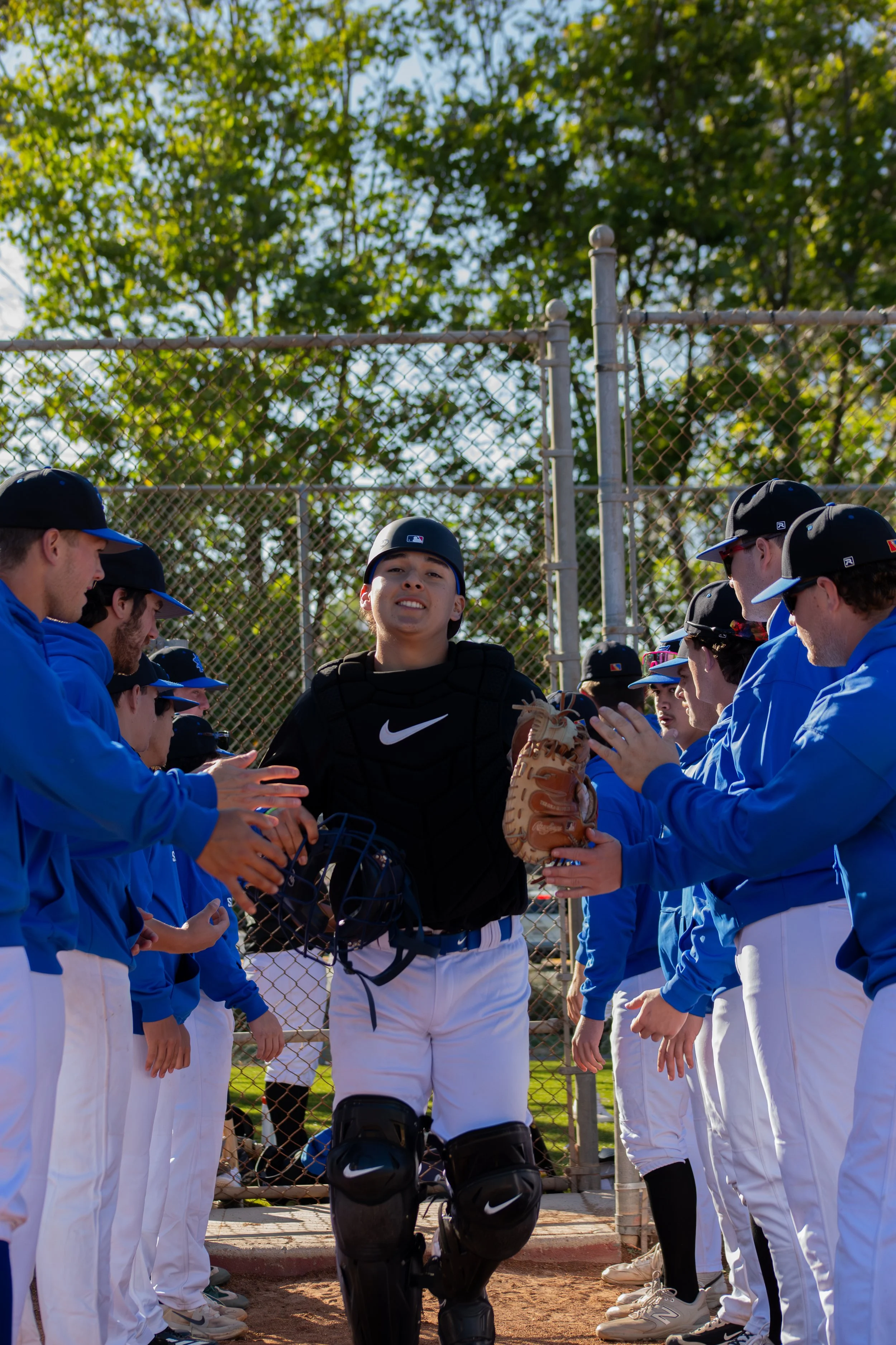 Youth baseball players in blue uniforms and a catcher in black gear standing in formation around a young male player in a black shirt, holding a baseball glove, near a chain-link fence with trees in the background.