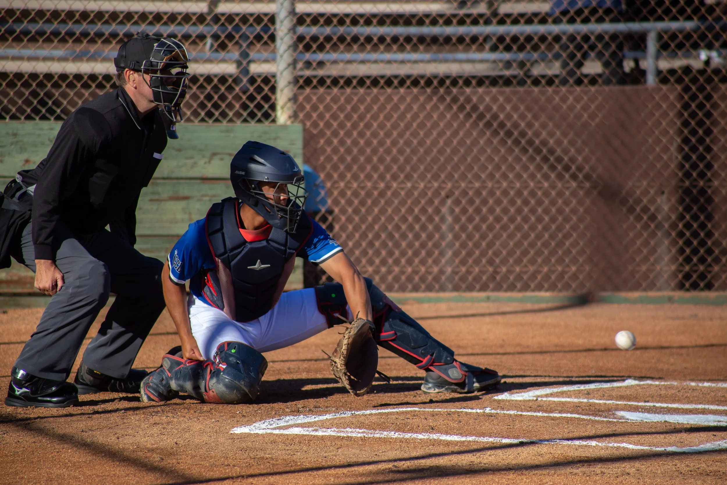 A baseball catcher in full gear getting ready to catch a pitch during a game. A baseball umpire stands behind him on a dirt field with a chain-link fence in the background.