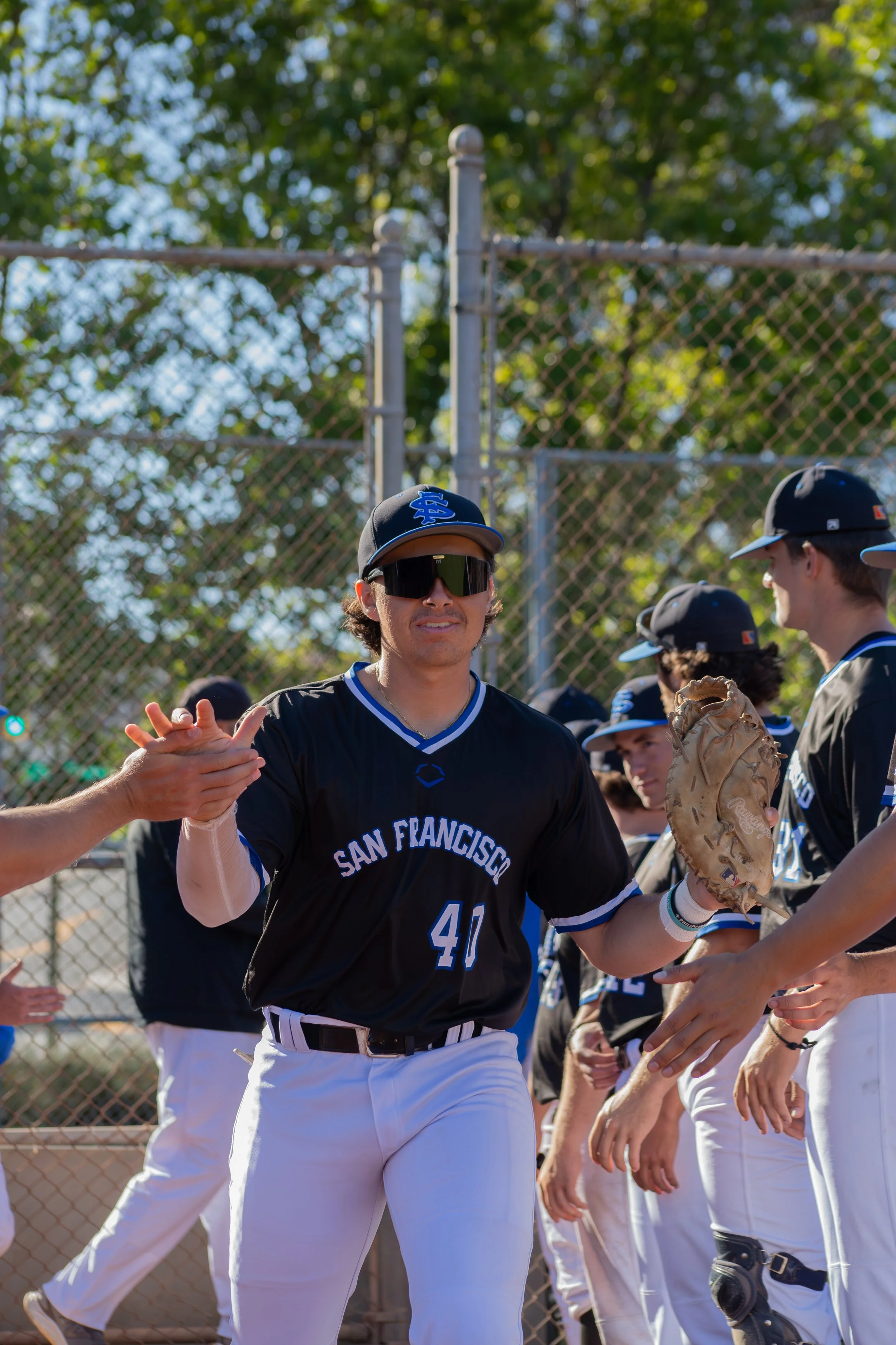 A baseball player from San Francisco, wearing sunglasses and a black jersey, shaking hands with teammates on the field with a chain-link fence and trees in the background.
