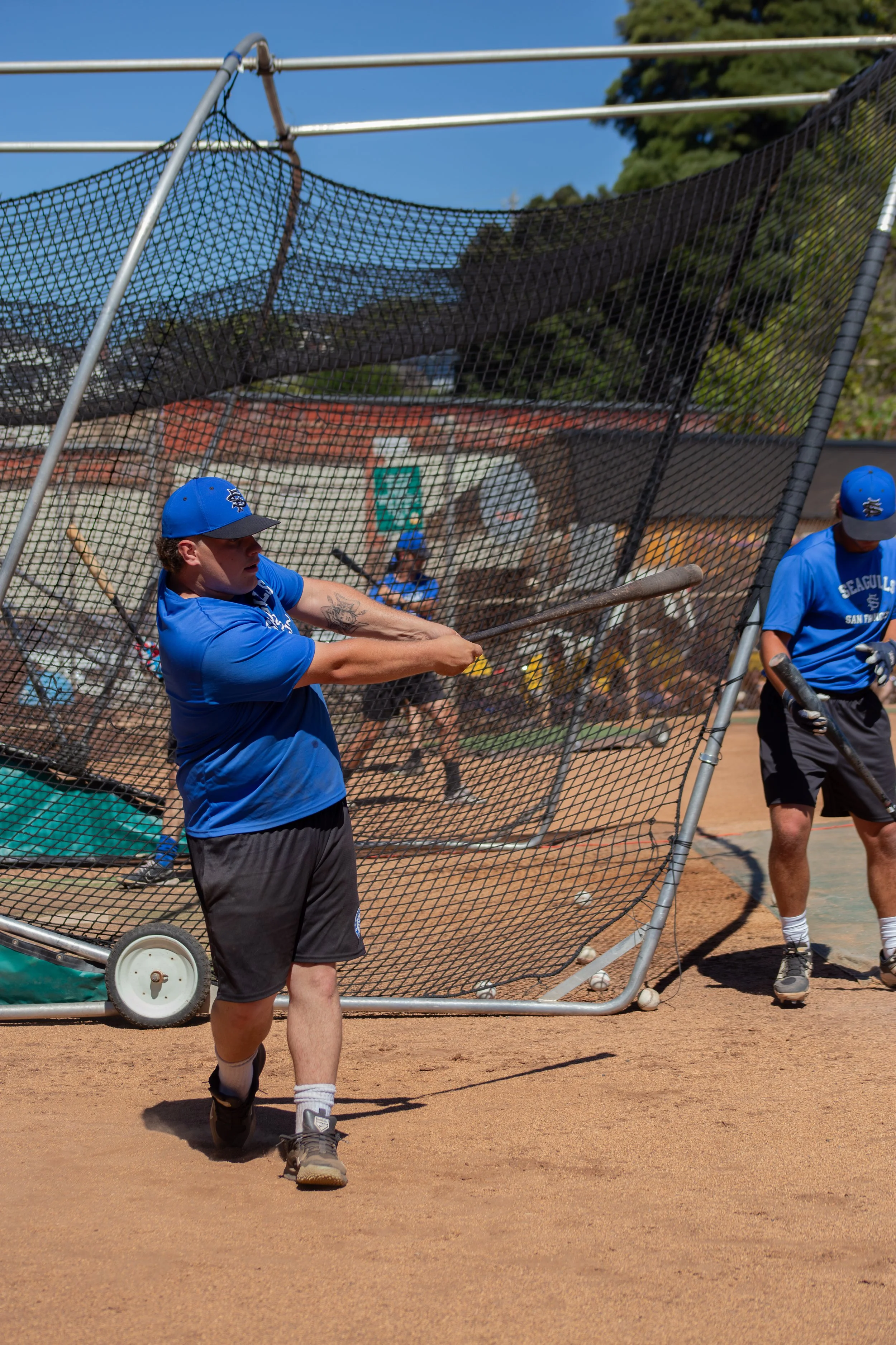 Young male baseball player in a blue uniform and cap practicing batting at practice cage on baseball field, with teammates and a dugout in the background.