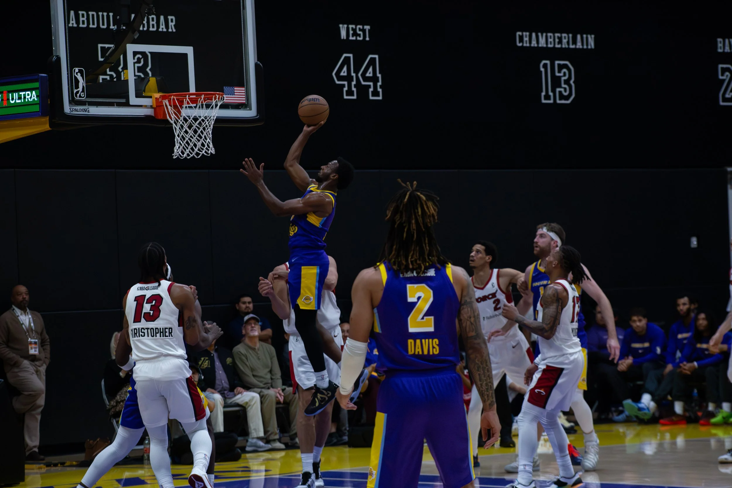 A basketball game action shot shows a player from the Golden State Warriors in blue and yellow uniform jumping to make a shot near the basket, with several players and spectators watching.