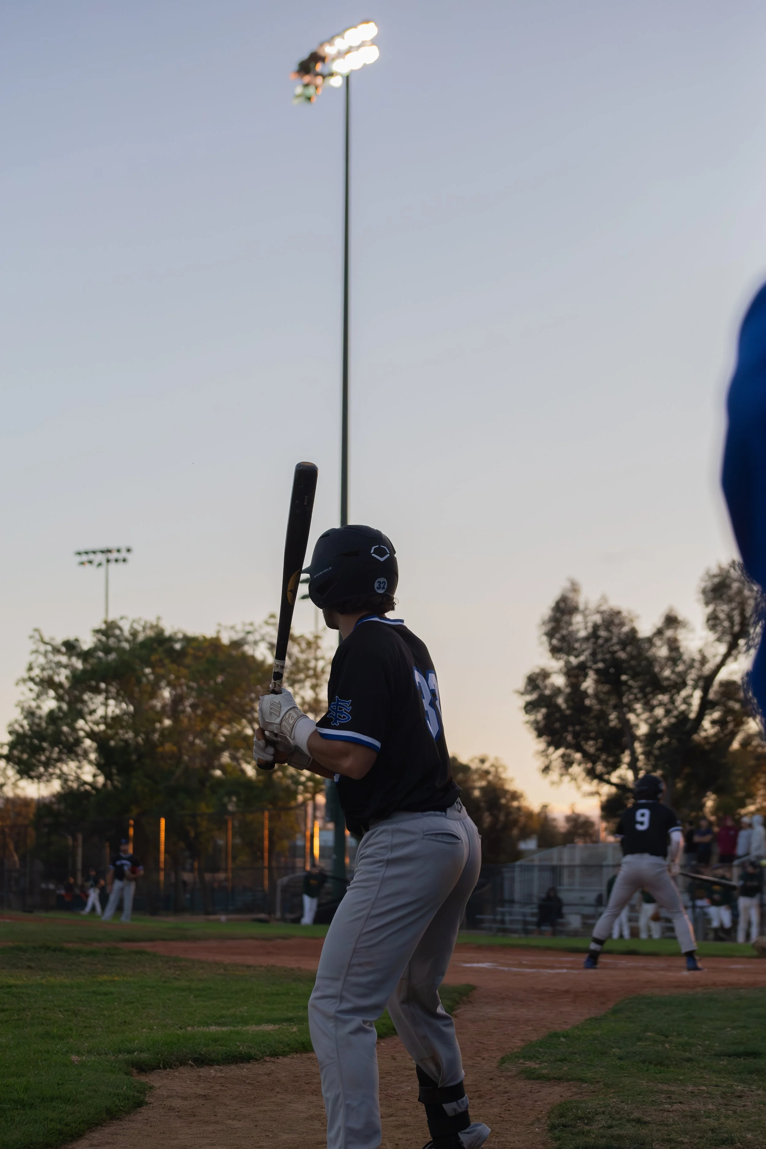 A baseball player in a black jersey and grey pants holding a bat at a baseball field during dusk. Other players are in the background, and stadium lights are visible in the sky.