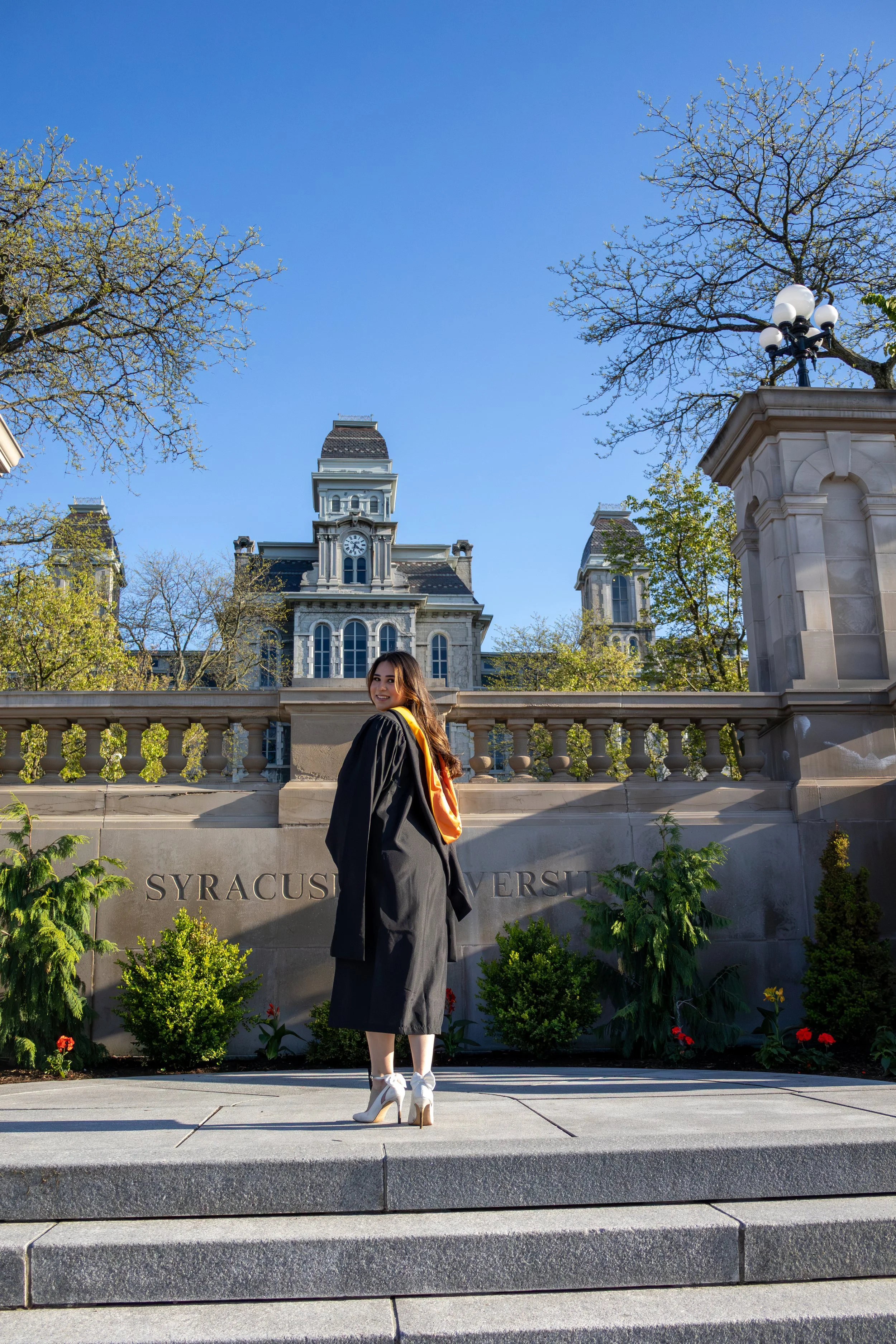 A young woman in graduation gown and heels standing on steps outside Syracuse University, with historic buildings and trees in the background.