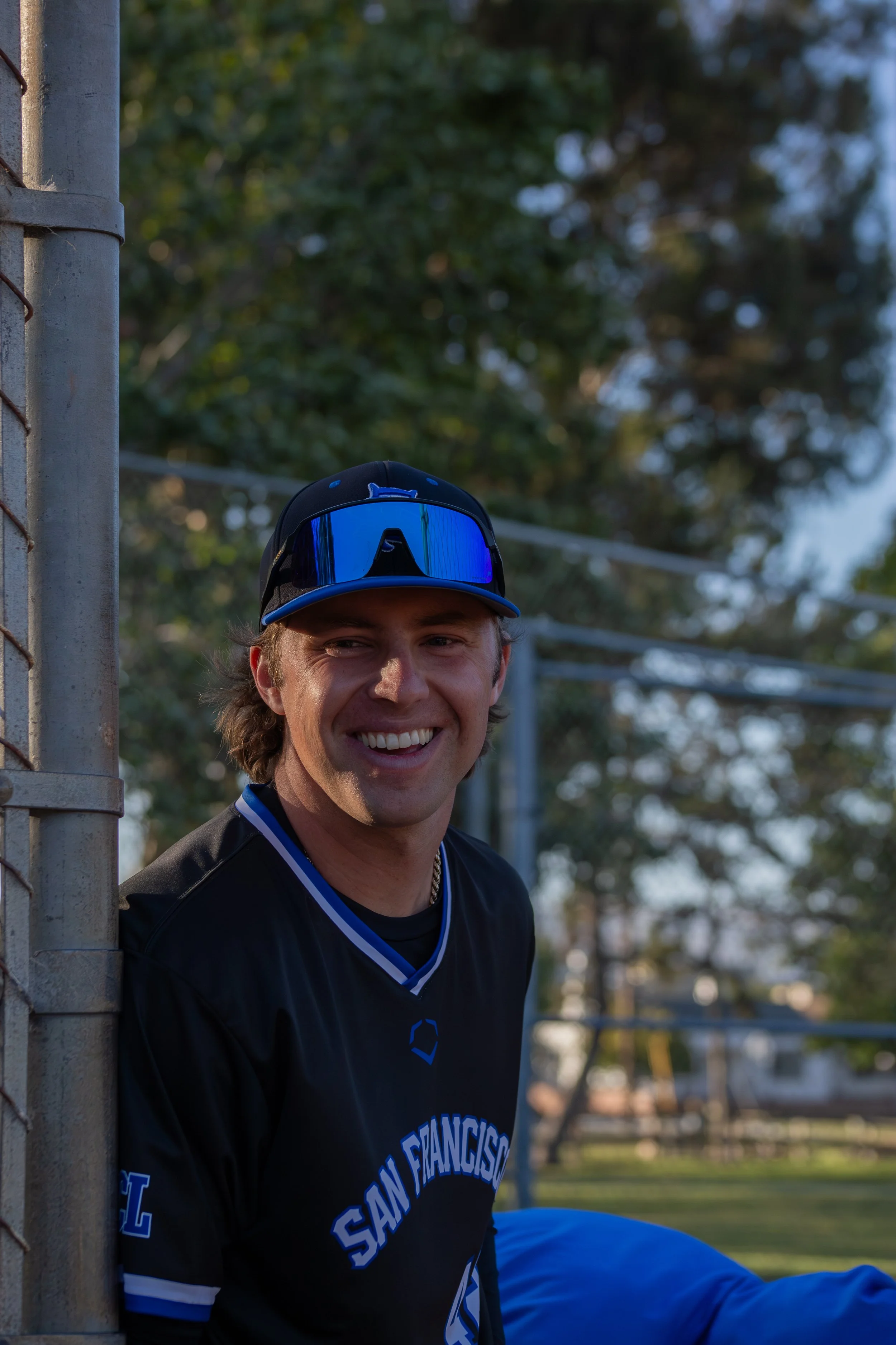 A young man smiling at the camera, wearing a black San Francisco Giants baseball jersey and a blue sports cap with sunglasses on top, standing next to a metal fence in a park with trees in the background.
