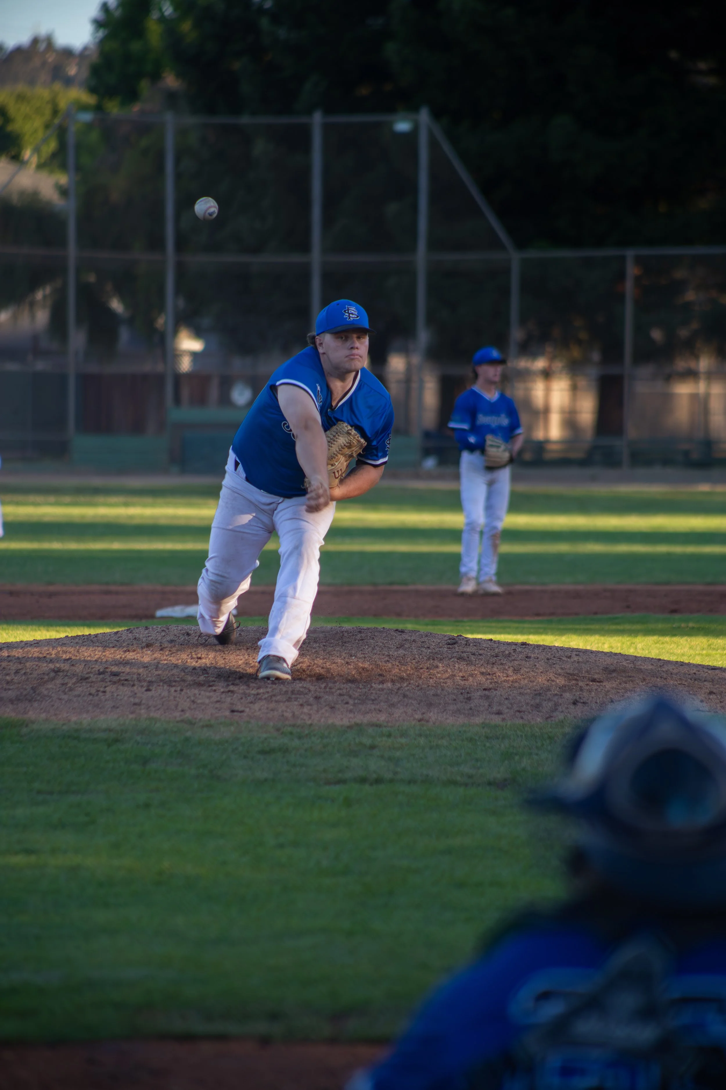 A baseball player in a blue uniform on a baseball field, preparing to pitch, with another player in the background and a baseball mid-air in front of him.
