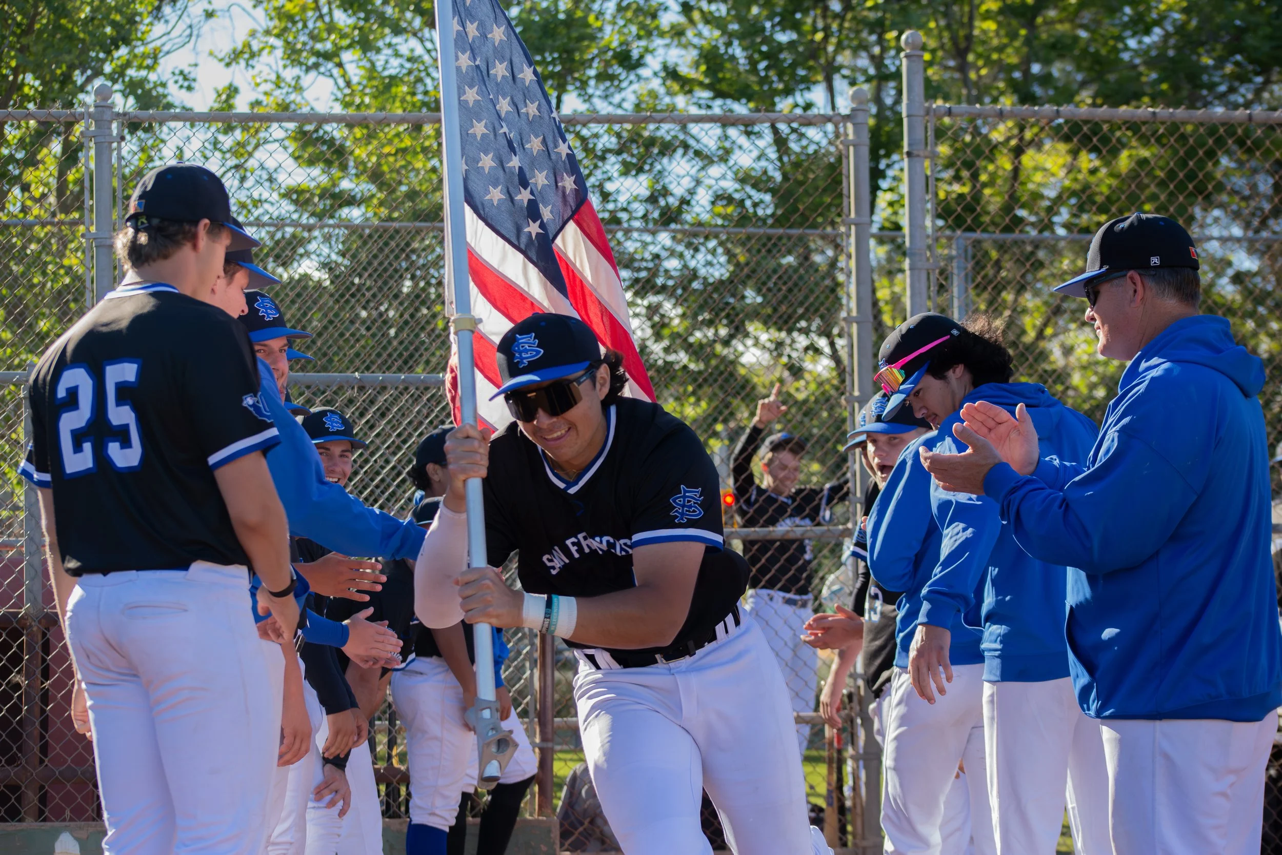 A group of baseball players and a coach are gathered around a flagpole with an American flag during a game. The players are wearing blue and white uniforms and black caps, and they appear to be celebrating or congratulating each other.