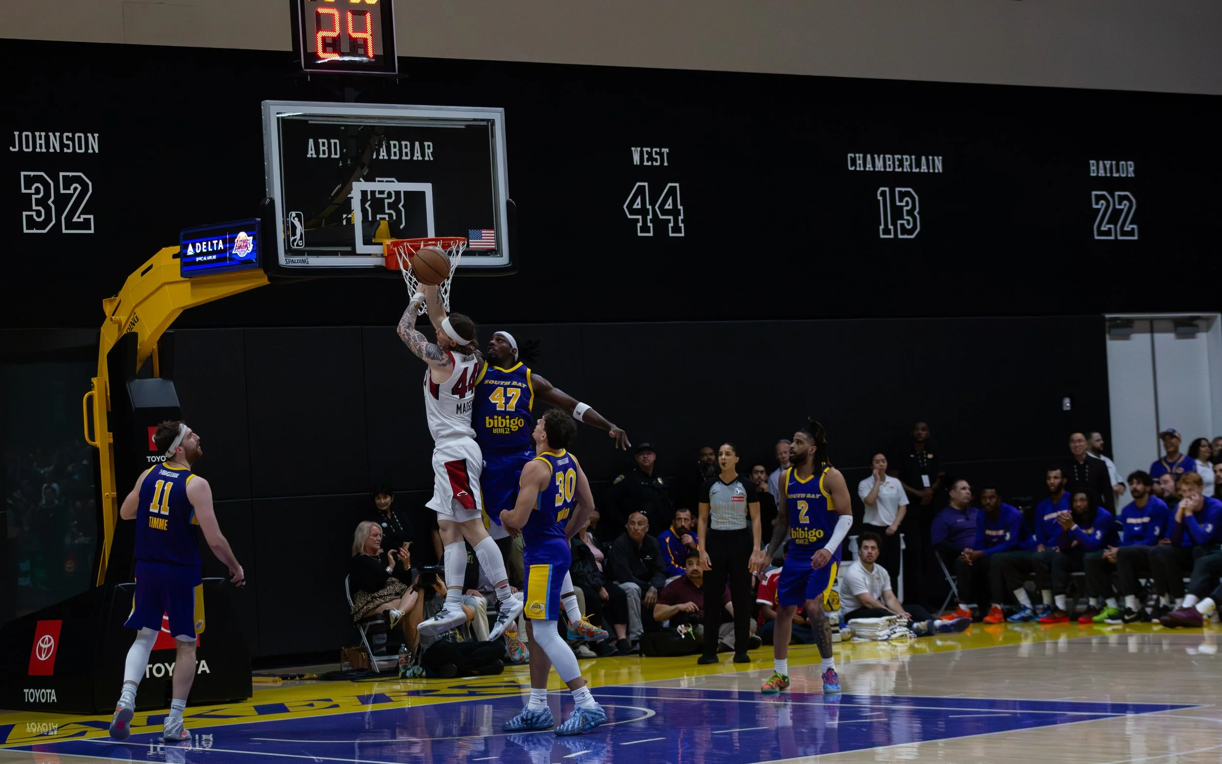 A basketball player in a white uniform jumping to shoot near the basket defended by a player in a blue uniform. Several players and spectators are visible around the court.