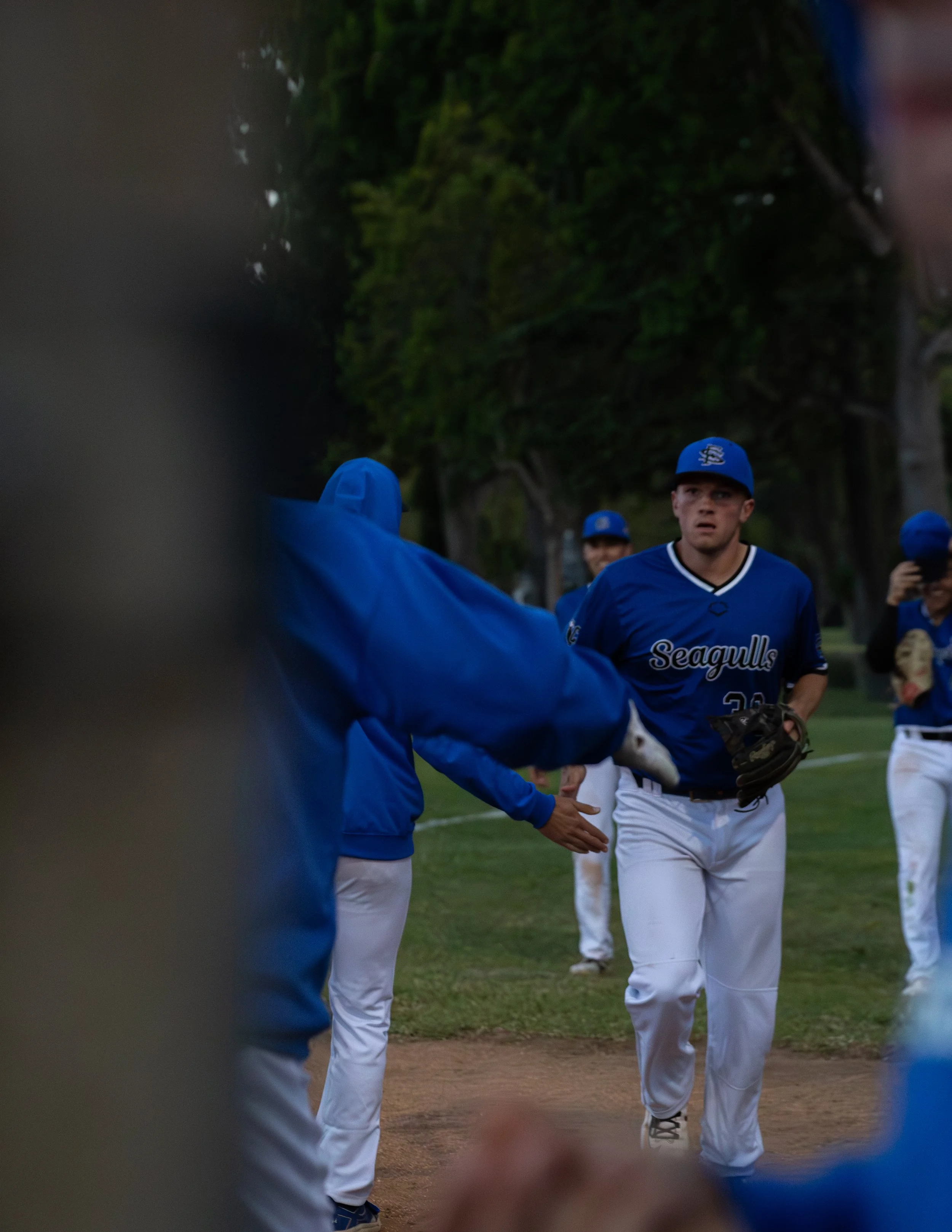 A baseball player wearing a blue uniform with 'Seagulls' written on it, standing on a field during a game, with trees in the background. The photo is partially obstructed by a blurred object in the foreground.