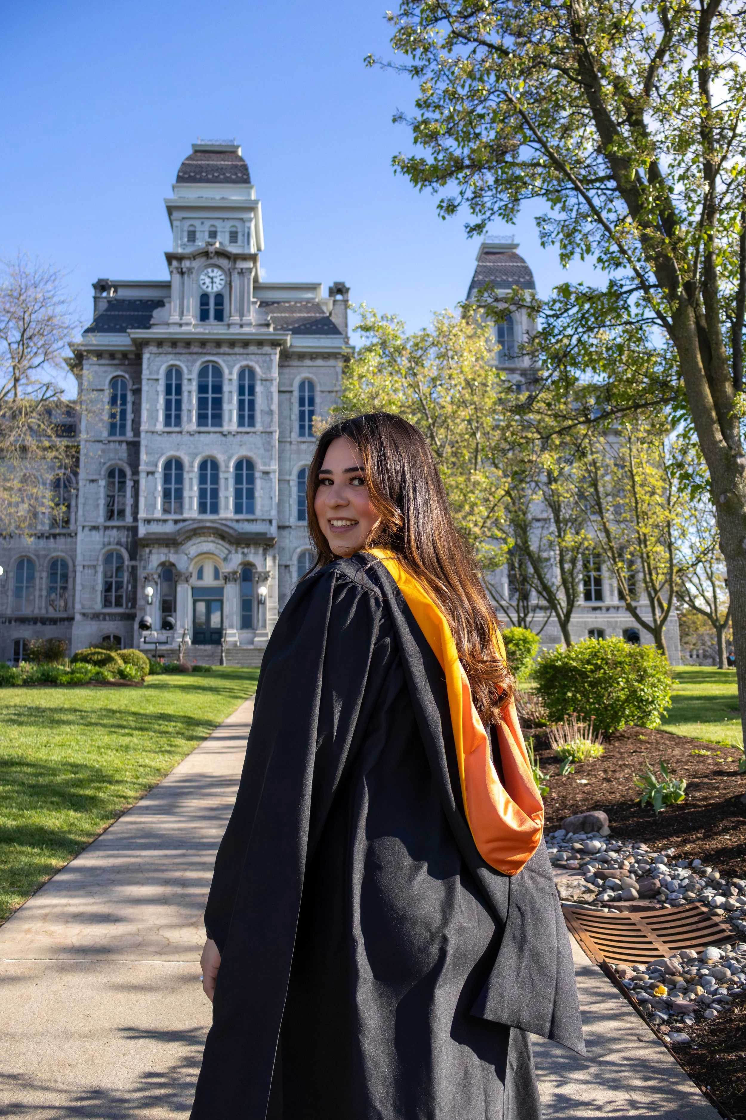 A young woman in a graduation gown and orange sash smiling outdoors, with a historic courthouse building in the background, surrounded by trees and a well-maintained garden on a sunny day.