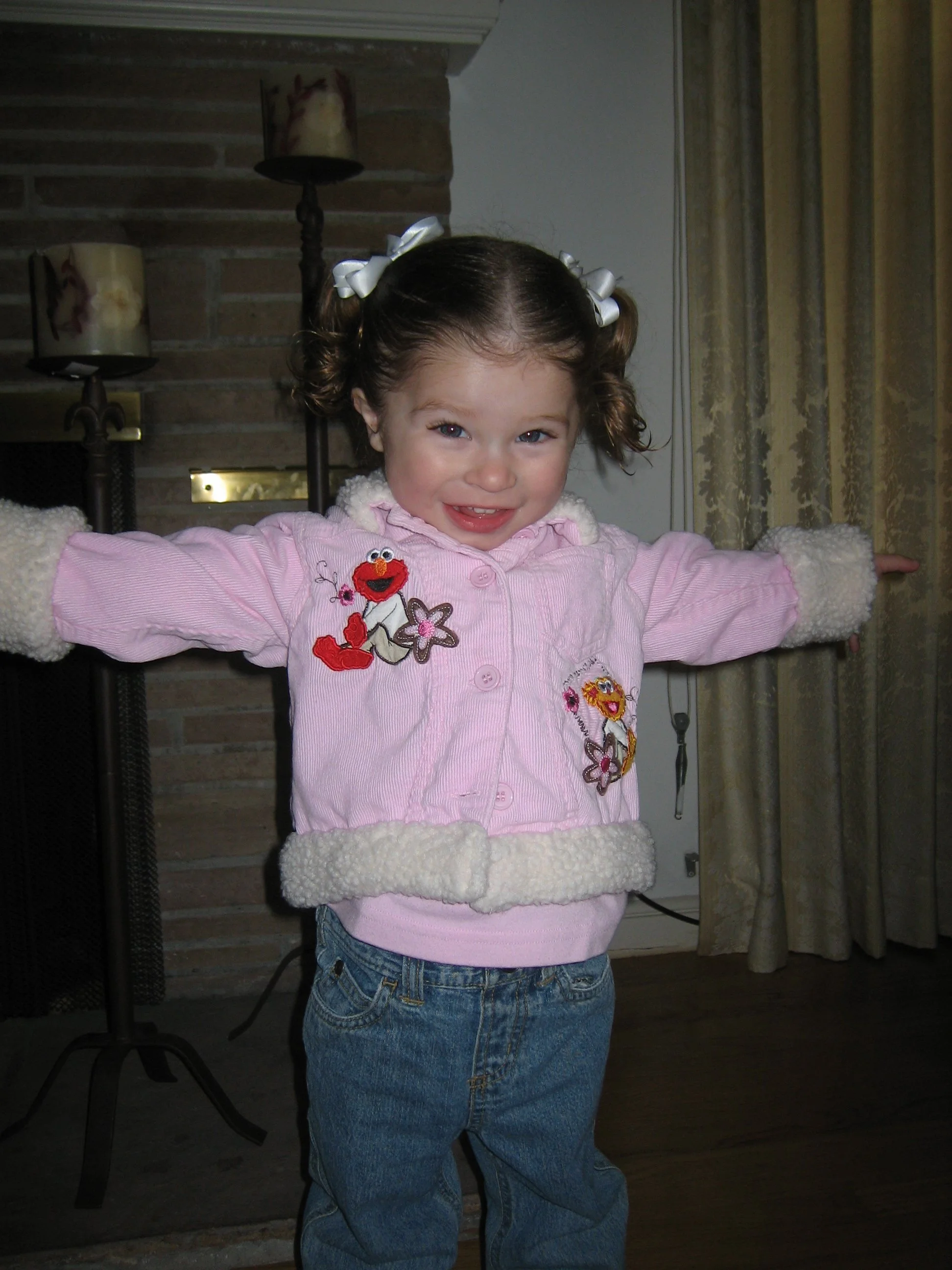 A young girl with brown hair styled in two pigtails with white ribbons, wearing a pink jacket with Sesame Street characters, and blue jeans, standing indoors with arms outstretched and smiling.