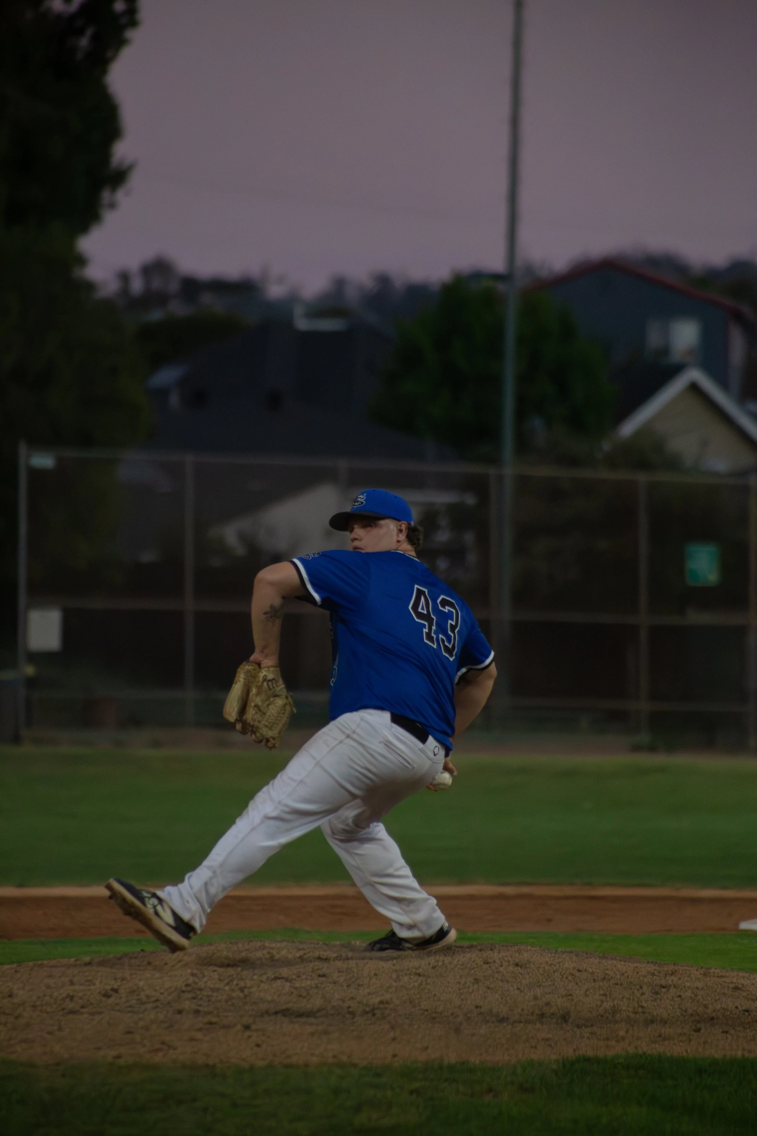 A baseball pitcher in a blue jersey with the number 43 on the back, wearing a cap, throwing a pitch on the mound during a game at dusk or early evening on a baseball field with houses and trees in the background.