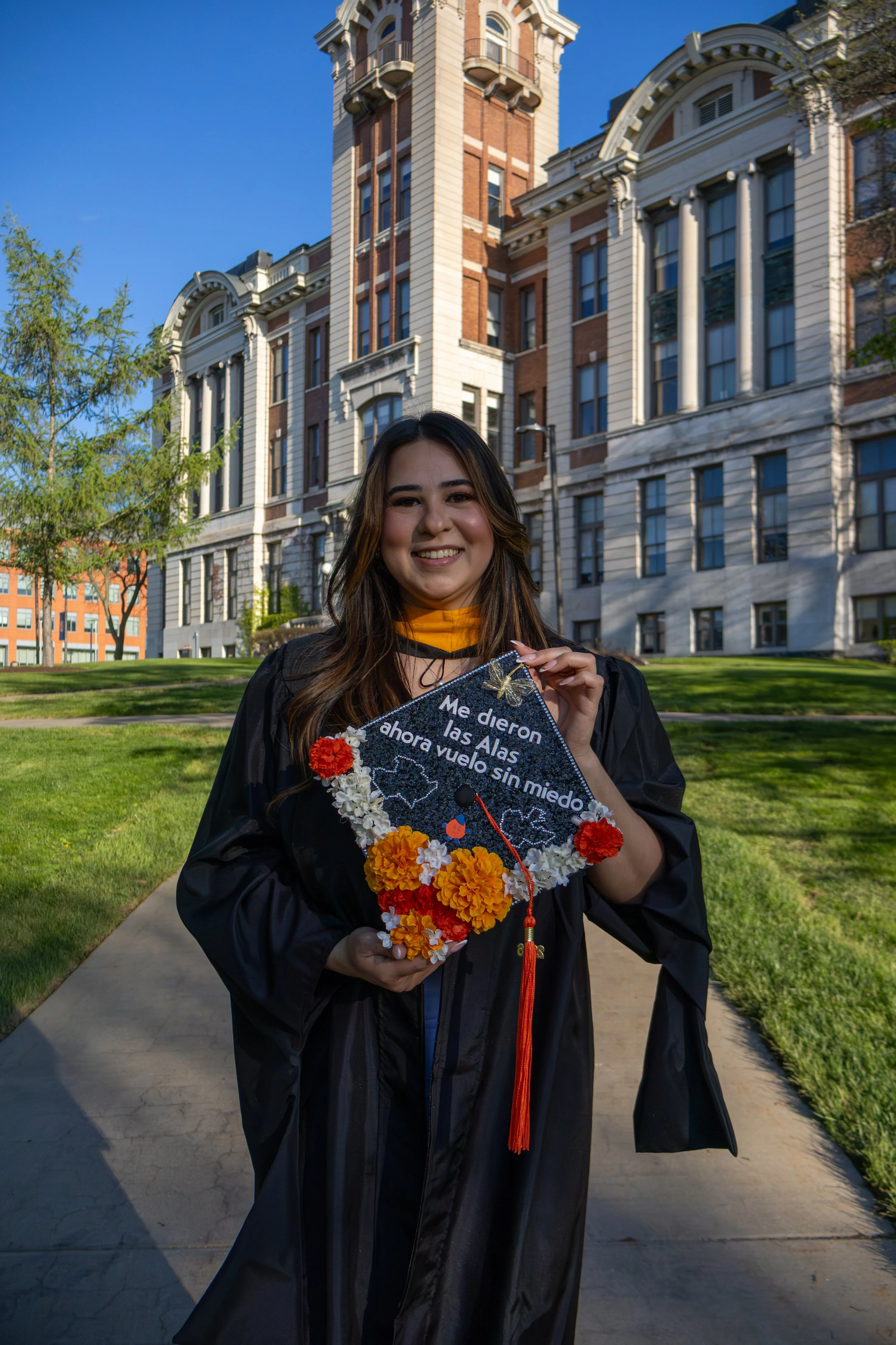 A young woman in a graduation gown holding a decorated graduation cap with the phrase in Spanish, 'Me dieron las Alas y ahora vuelo sin miedo,' standing outdoors on a sunny day in front of a large historic building with a tower.