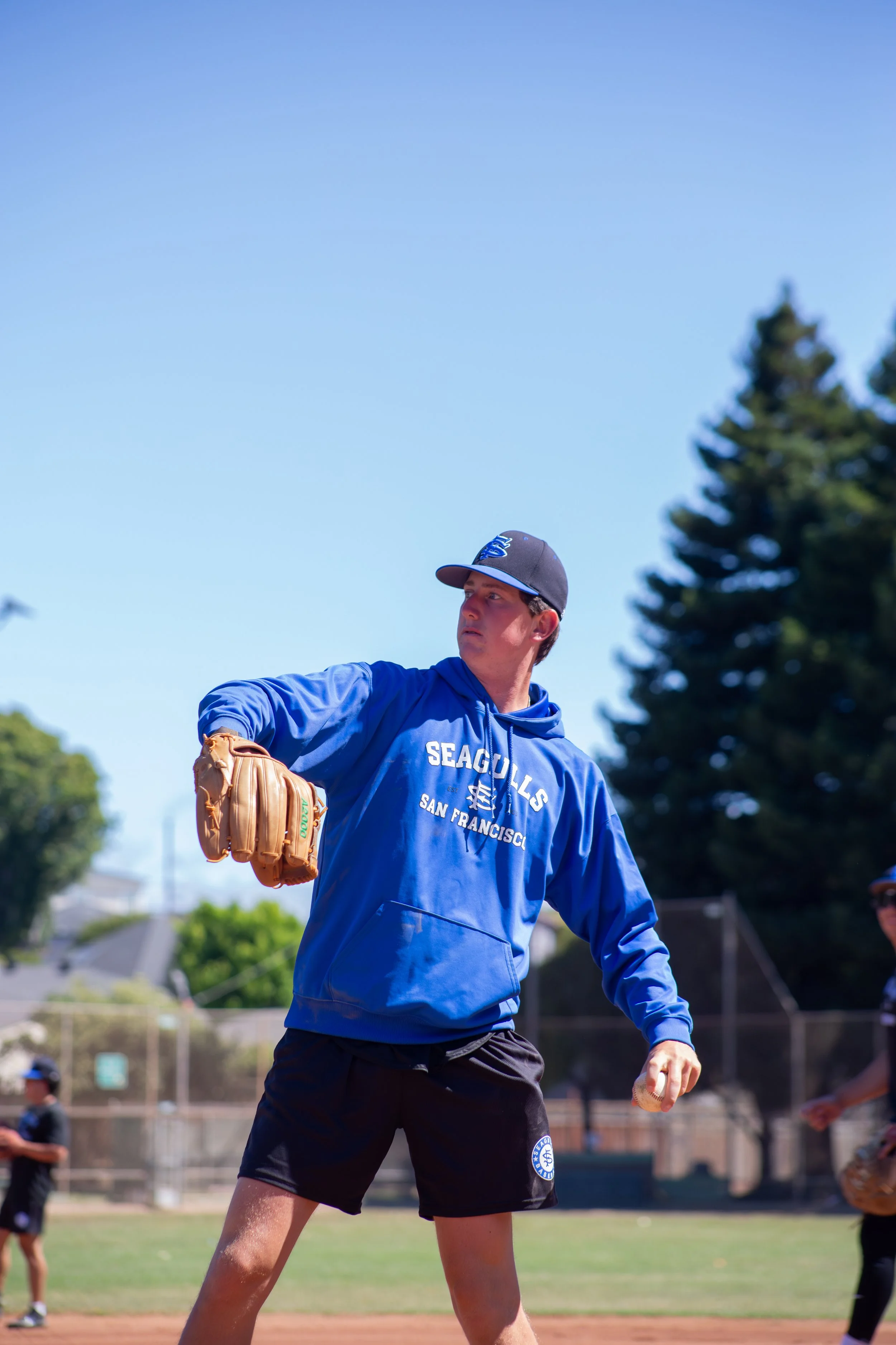 A young male baseball player in a blue hoodie and black shorts preparing to throw a baseball during a game on a sunny day, with other players and trees visible in the background.