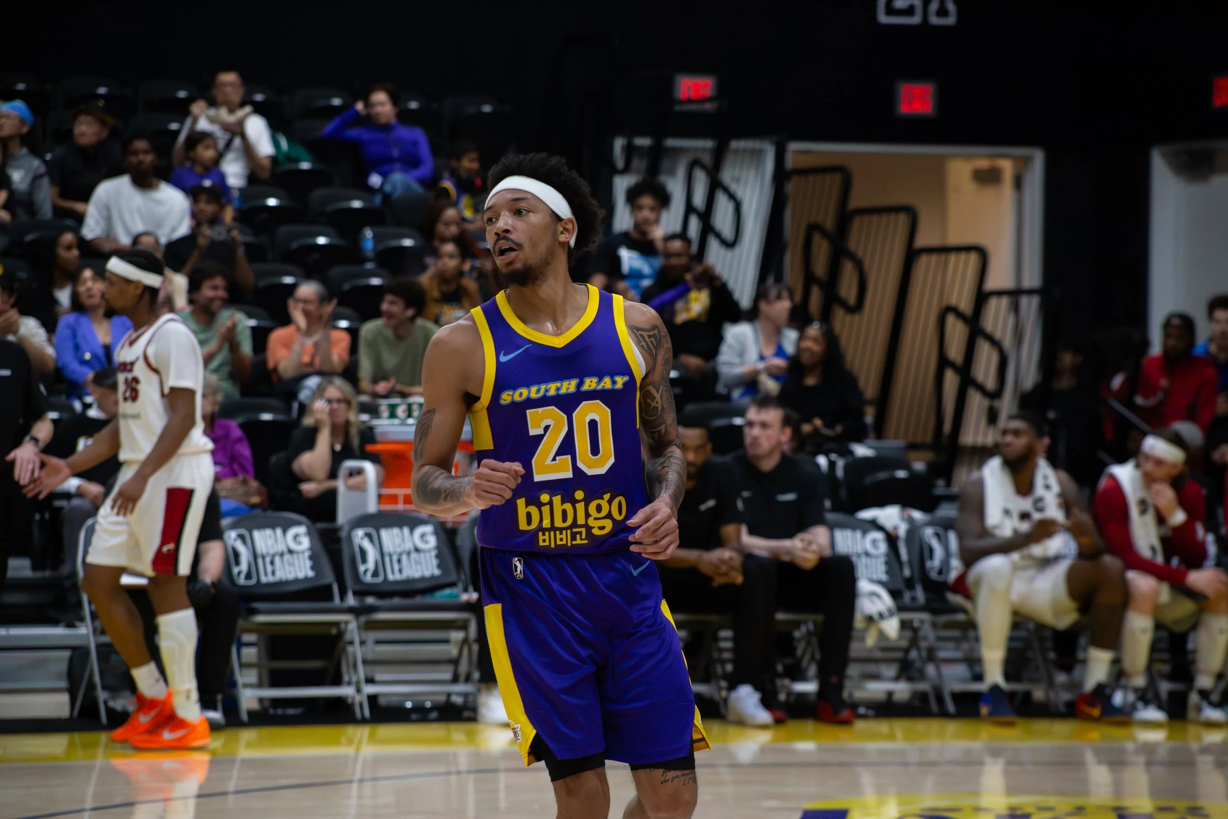 A basketball player wearing a blue and yellow jersey with the number 20, 'South Bay' written on it, and a white headband, standing on a basketball court during a game. In the background, there are spectators seated in black chairs, some clapping and 