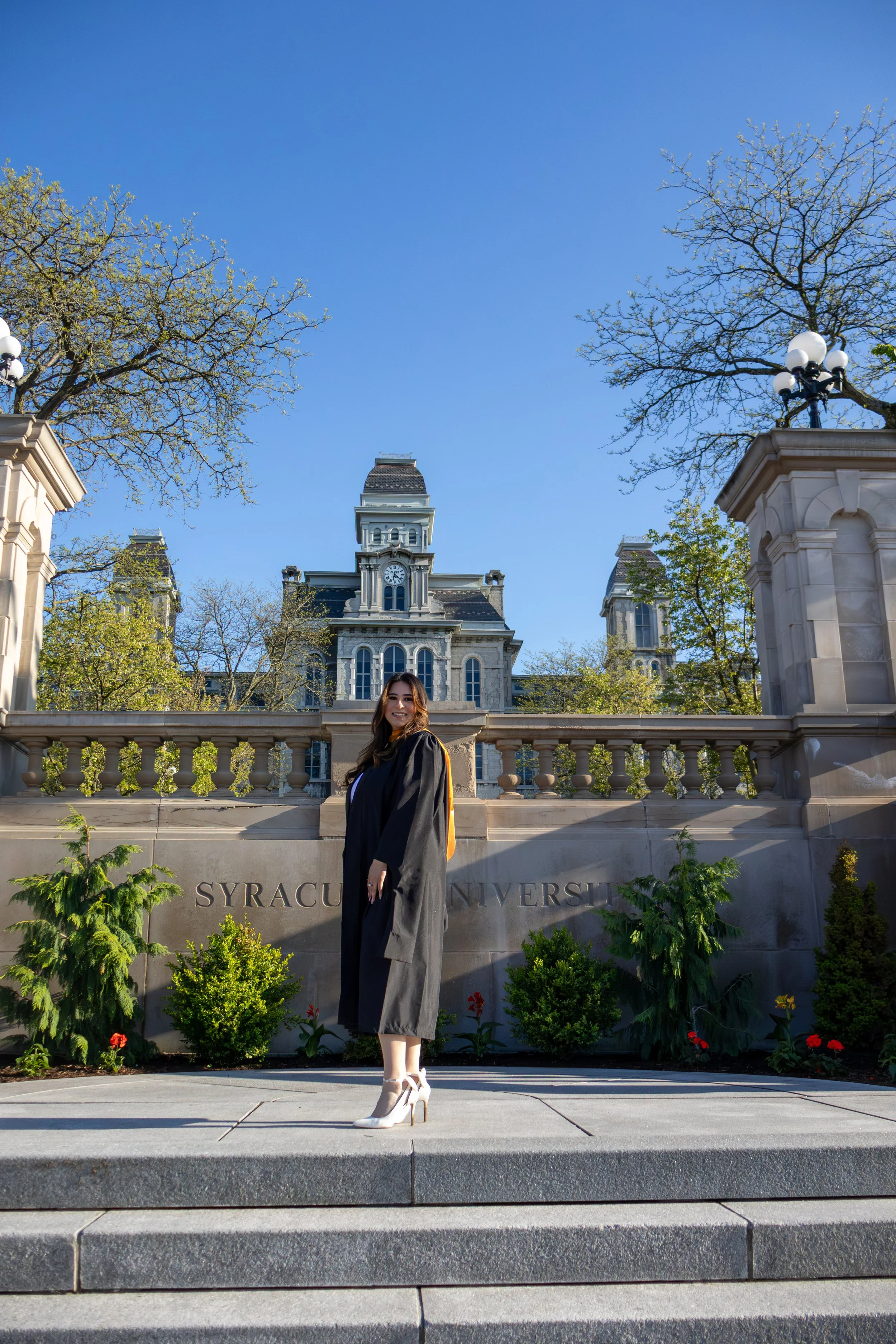 A woman in a graduation gown standing on steps in front of Syracuse University sign with a historic building and trees in the background.