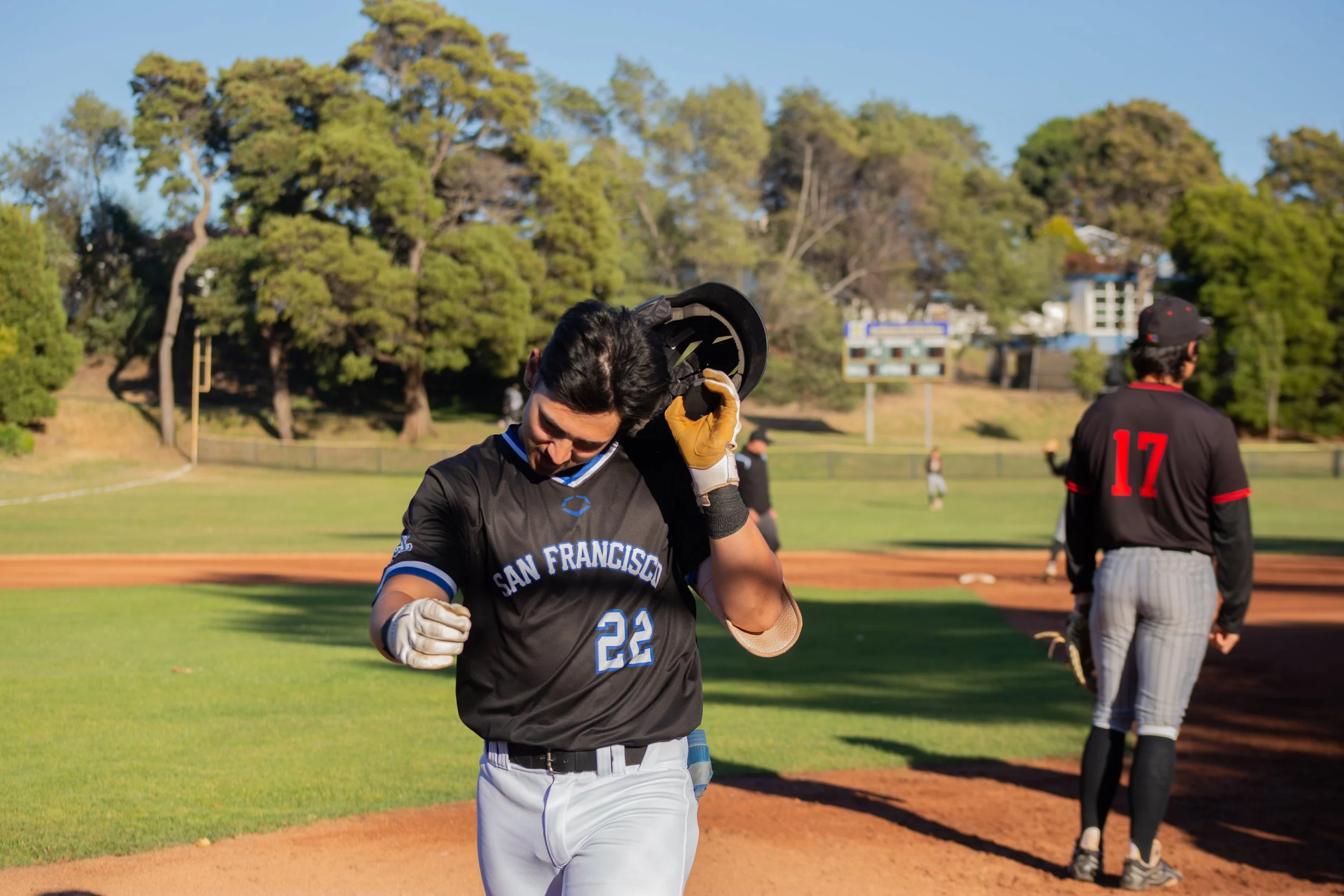 A baseball player from the San Francisco team putting on a helmet on a baseball field. Other players and trees are visible in the background during daytime.