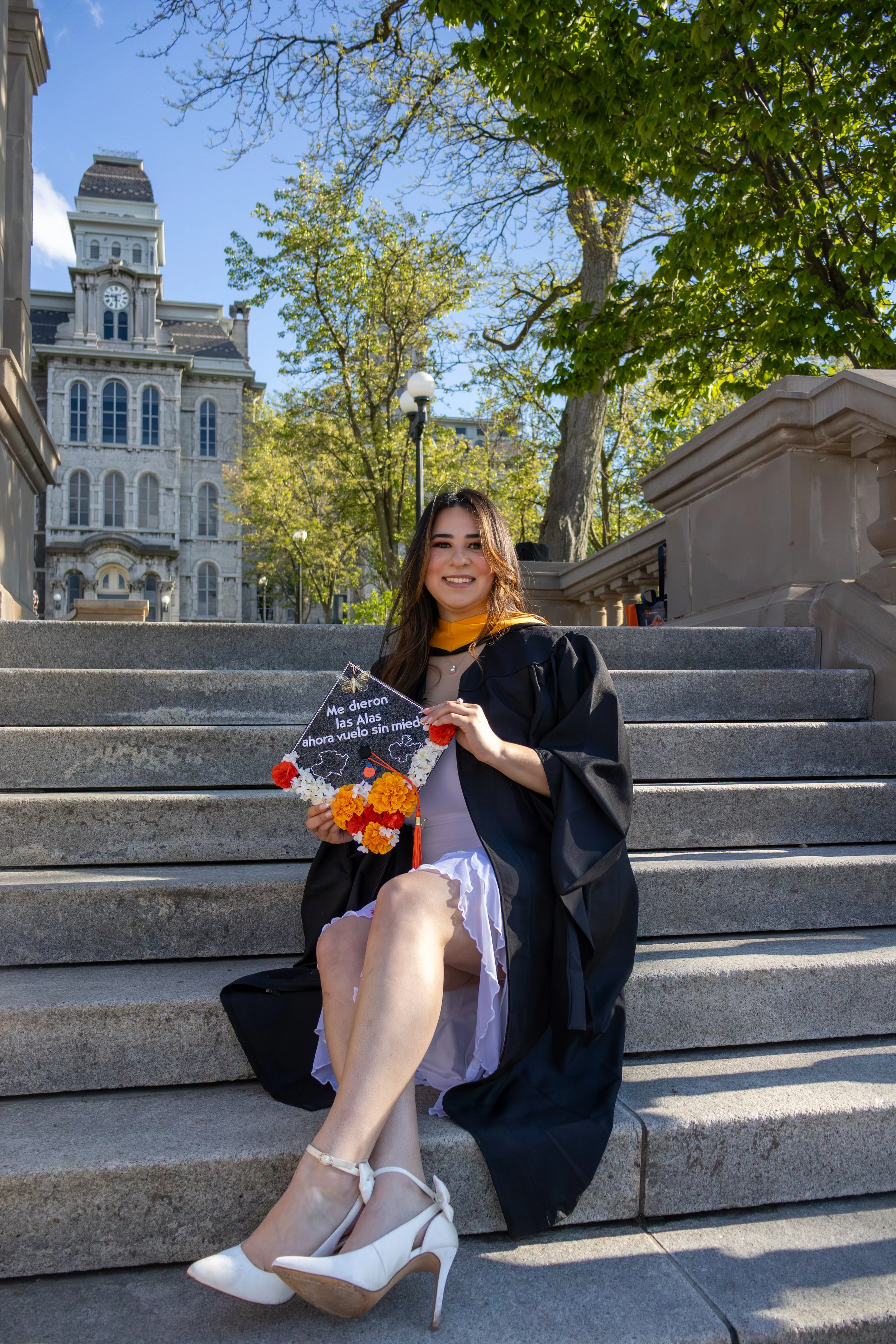 Young woman in graduation gown sitting on steps outdoors, holding a floral graduation cap with Spanish text, smiling, with historic building and trees in background.
