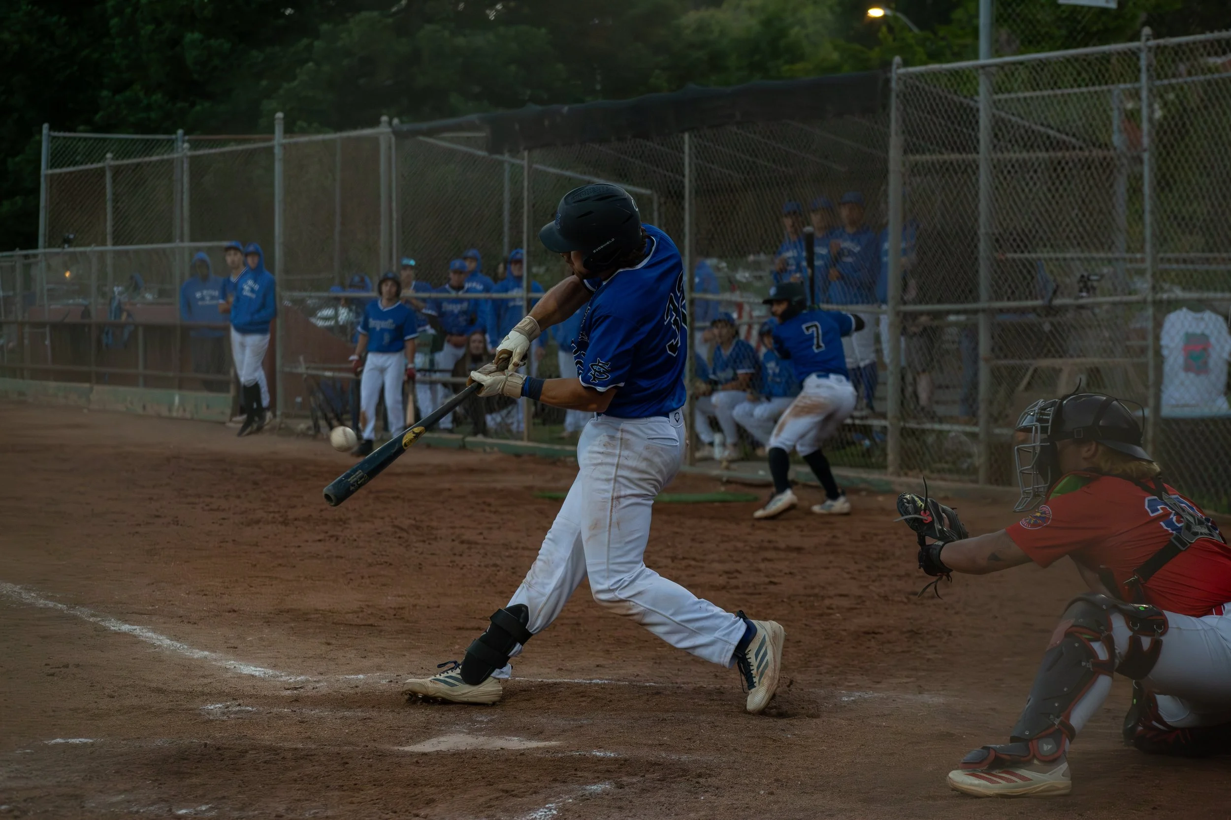 A baseball player in a blue jersey and white pants is swinging a bat at a pitched ball during a game on a dirt field. Other players and spectators, all in blue uniforms, are visible in the background behind a chain-link fence.
