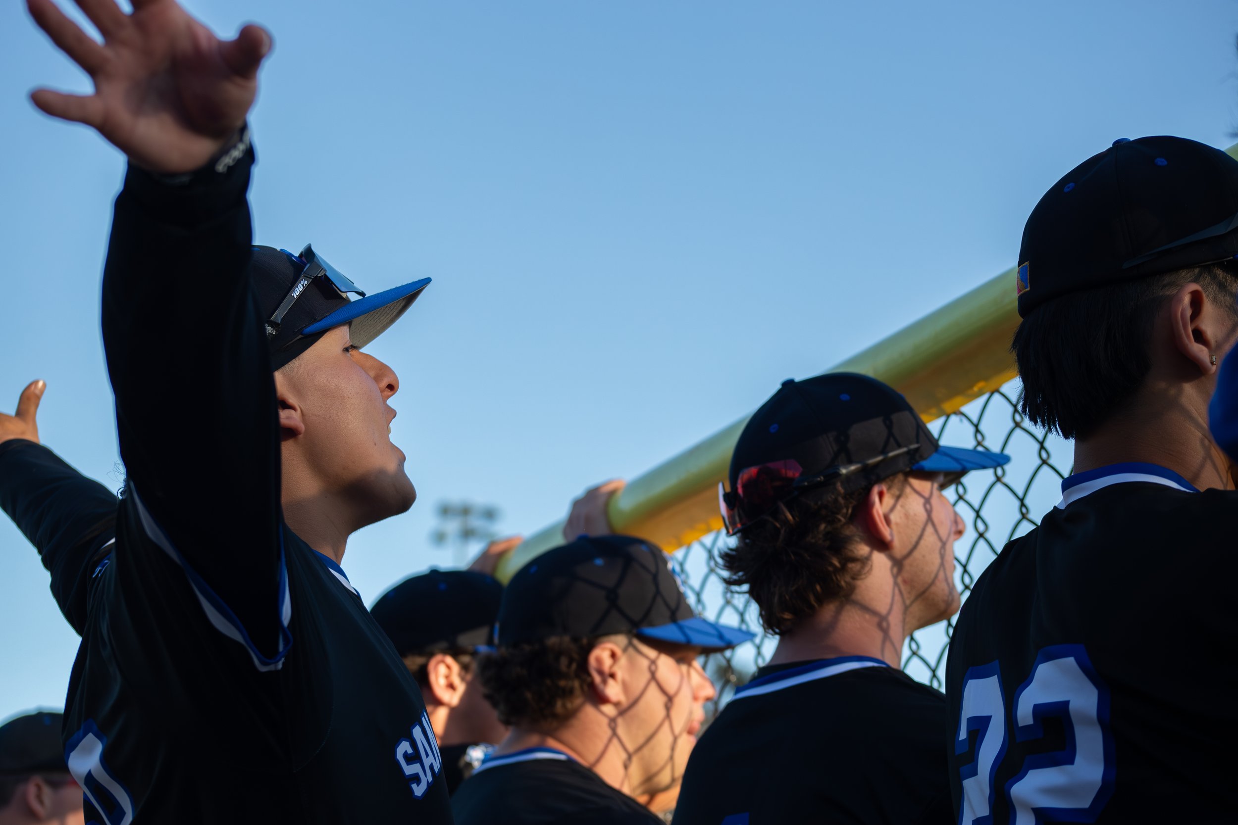 Group of softball players standing side by side with their hands on a yellow bat, wearing black uniforms and caps, as they look toward the field during a game under a blue sky.