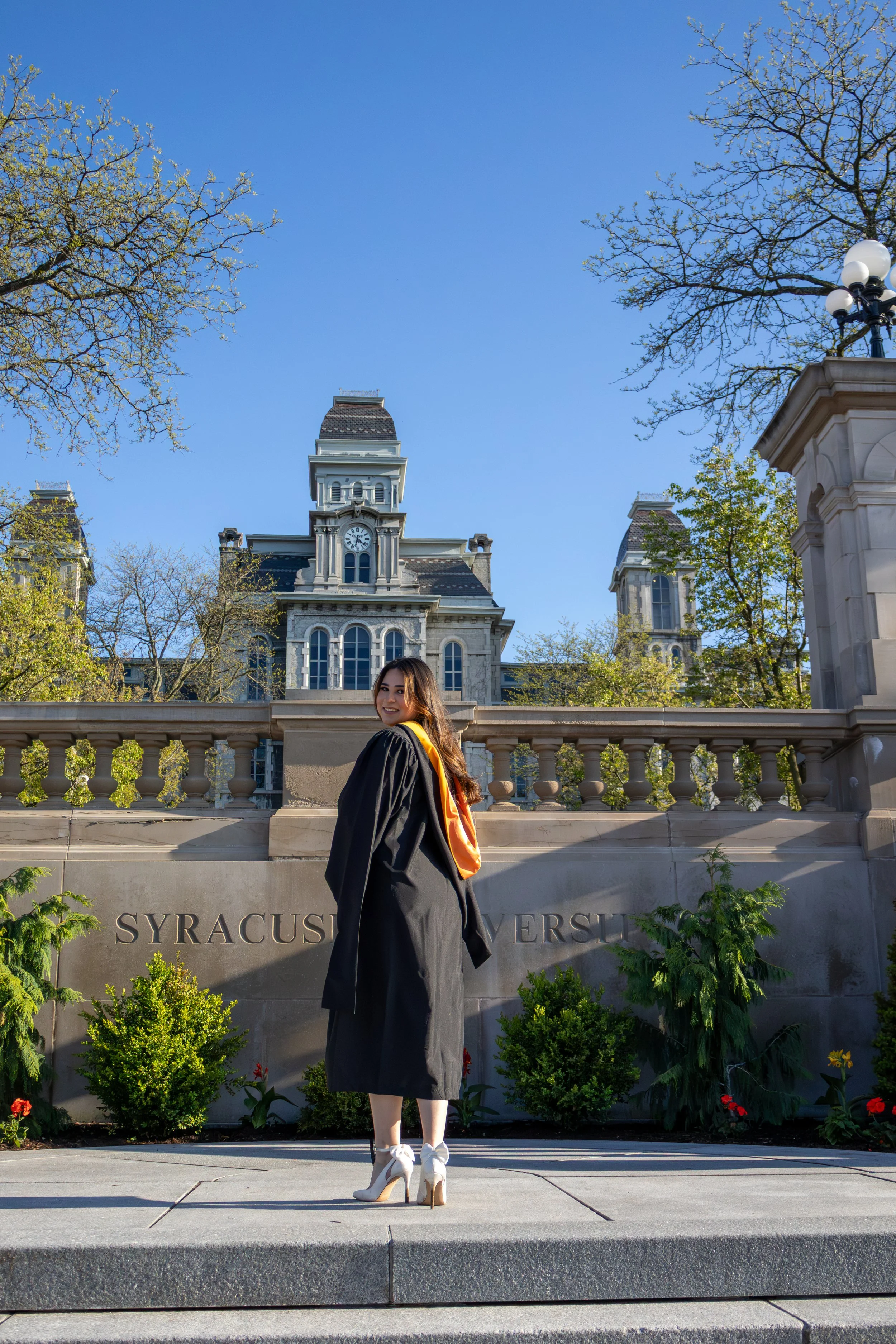 A young woman in a graduation gown and high heels posing outdoors in front of a stone wall with the words 'Syracuse University' engraved on it. The background shows a historic building with a clock tower, trees, and a clear blue sky.