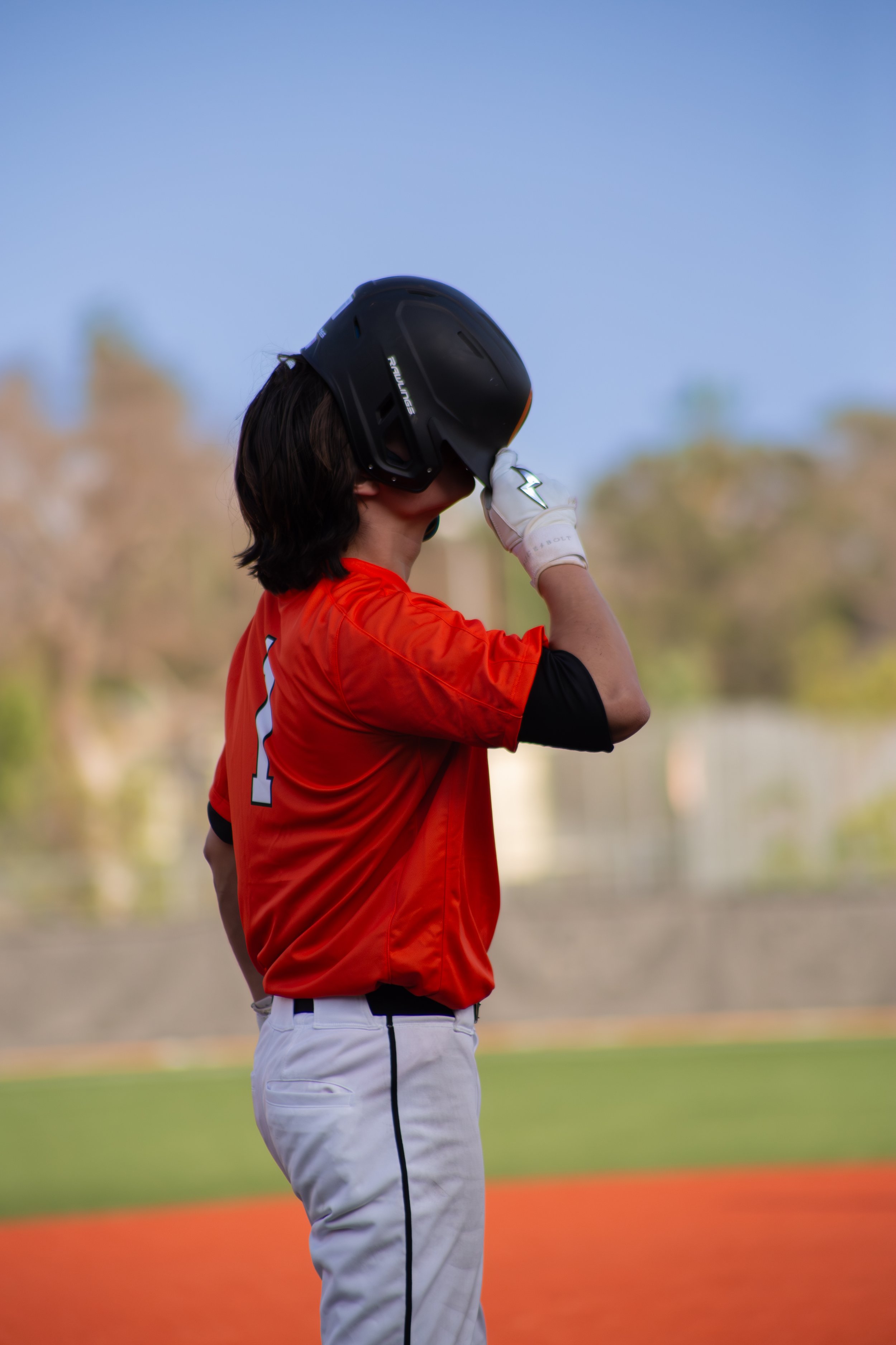 A person wearing a black helmet and an orange sports jersey, standing on a baseball field, adjusting the helmet.