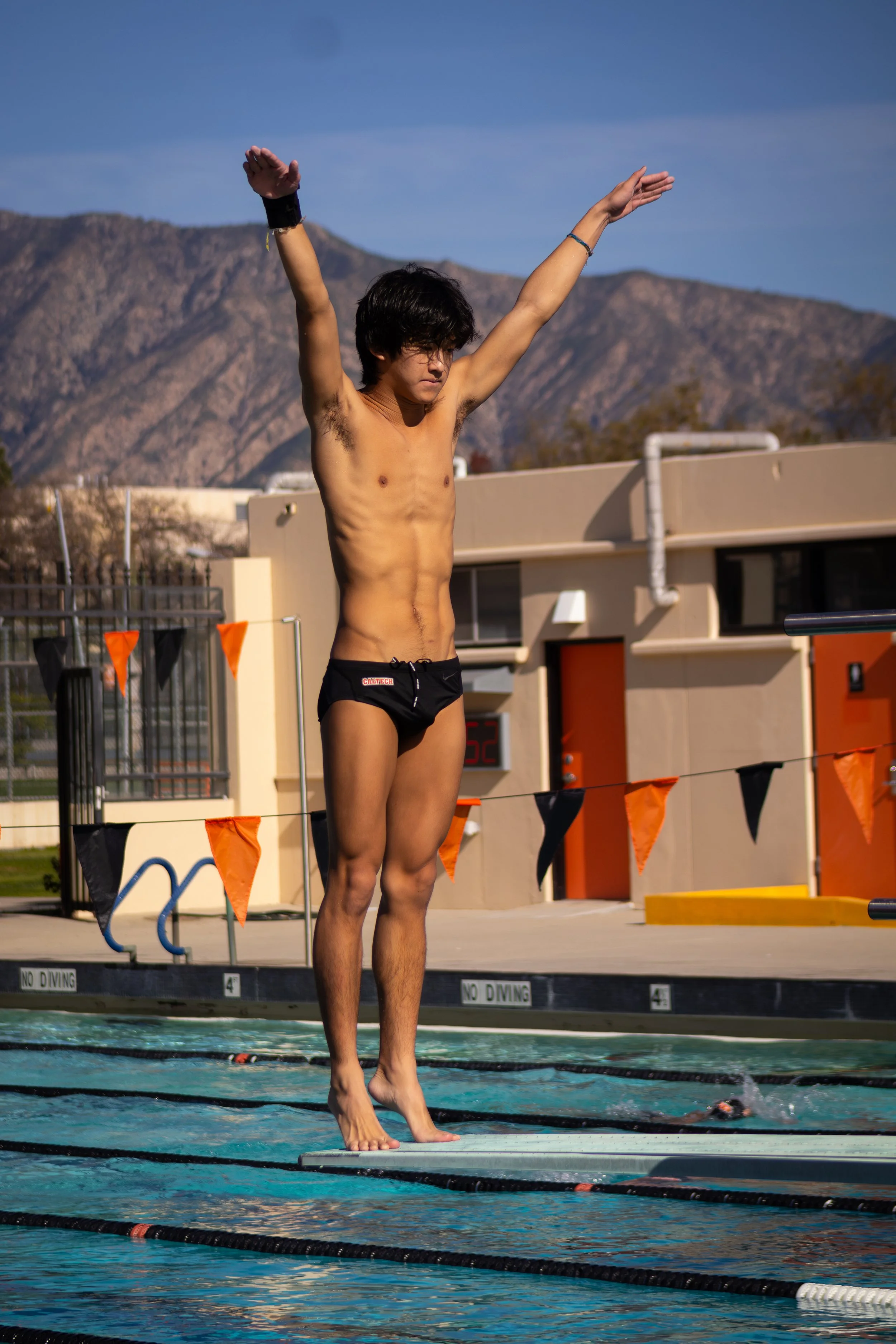 A male swimmer standing on a diving board at an outdoor pool with his arms raised, mountains and a building in the background.