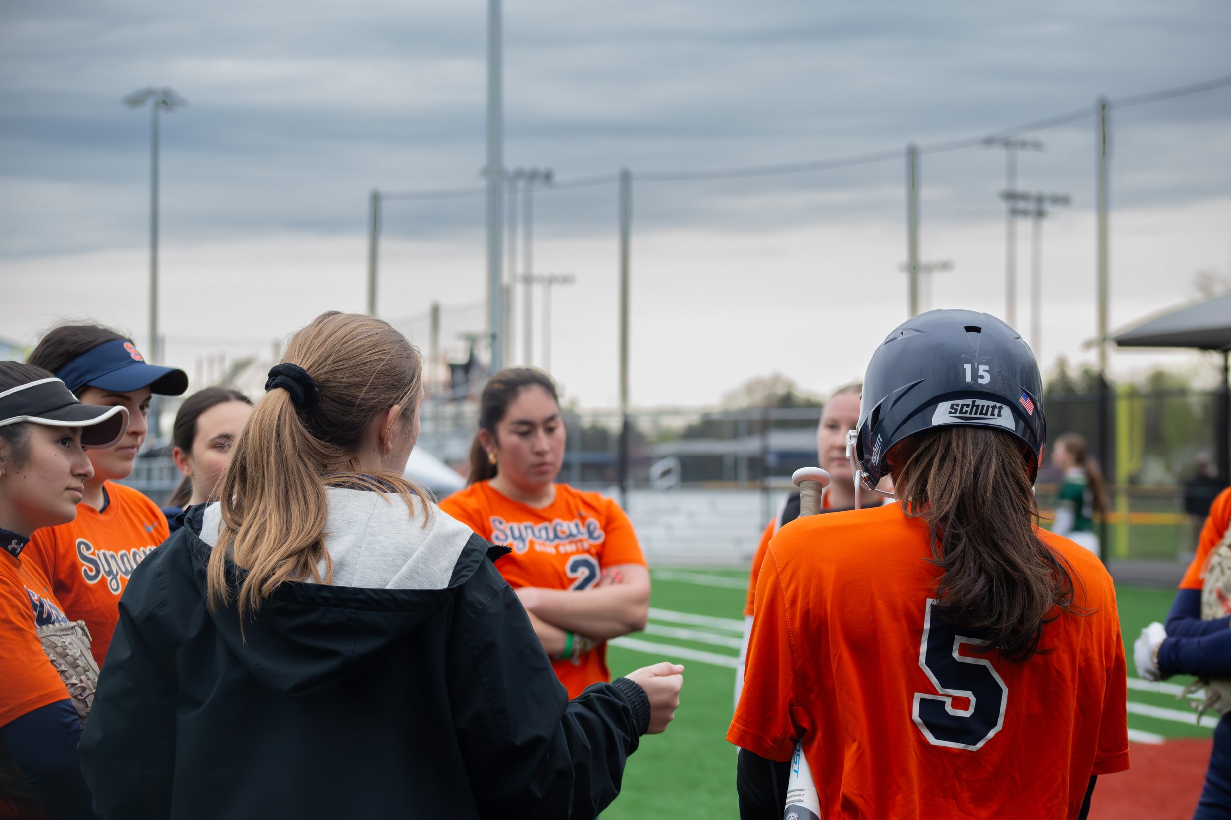 A group of young female athletes in orange jerseys gathered on a sports field, with some wearing helmets, in an outdoor stadium with a cloudy sky and athletic light poles in the background.
