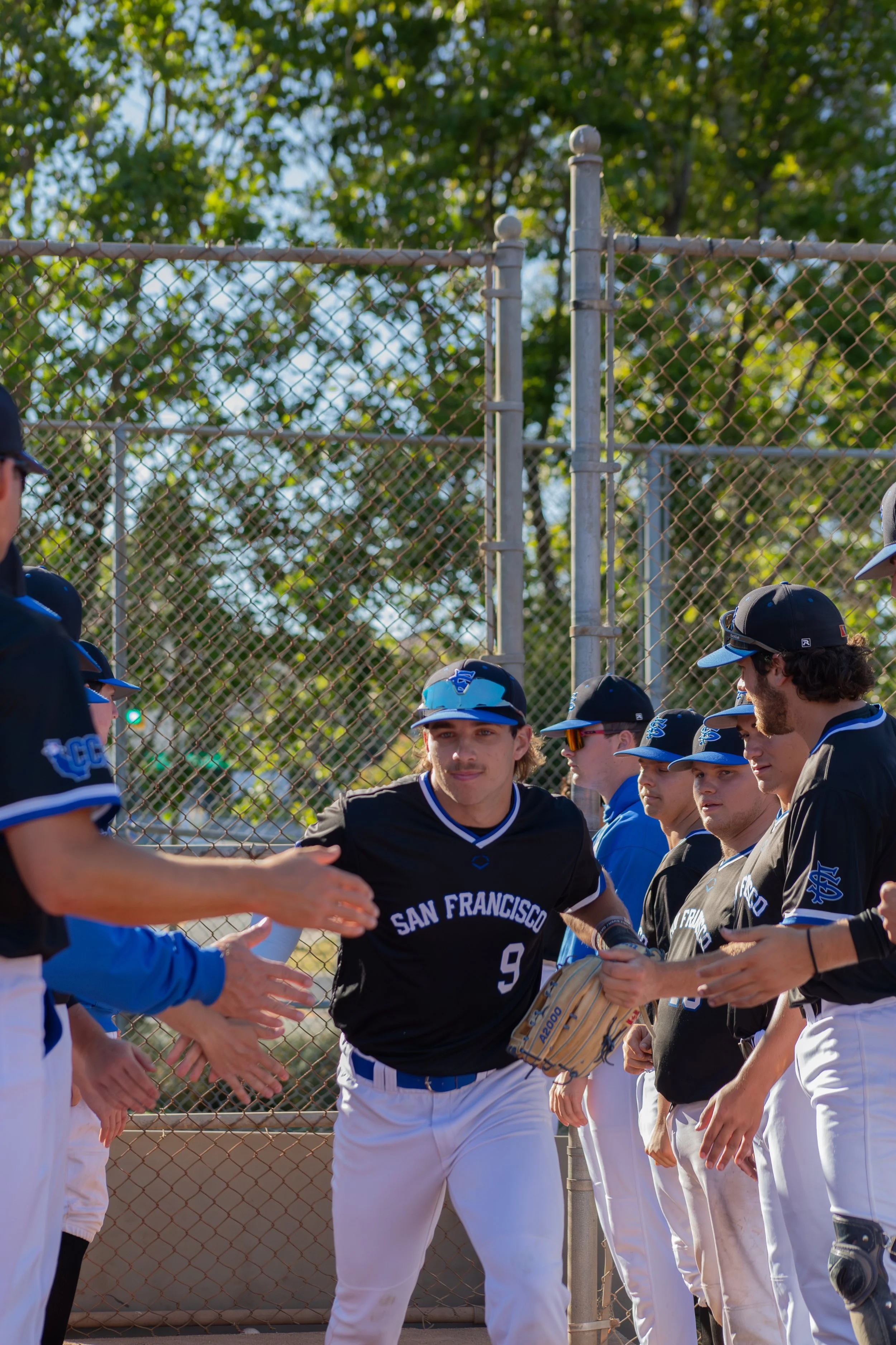Baseball players in black and blue uniforms shaking hands and exchanging high fives before a game at the baseball field, with a chain-link fence and trees in the background.