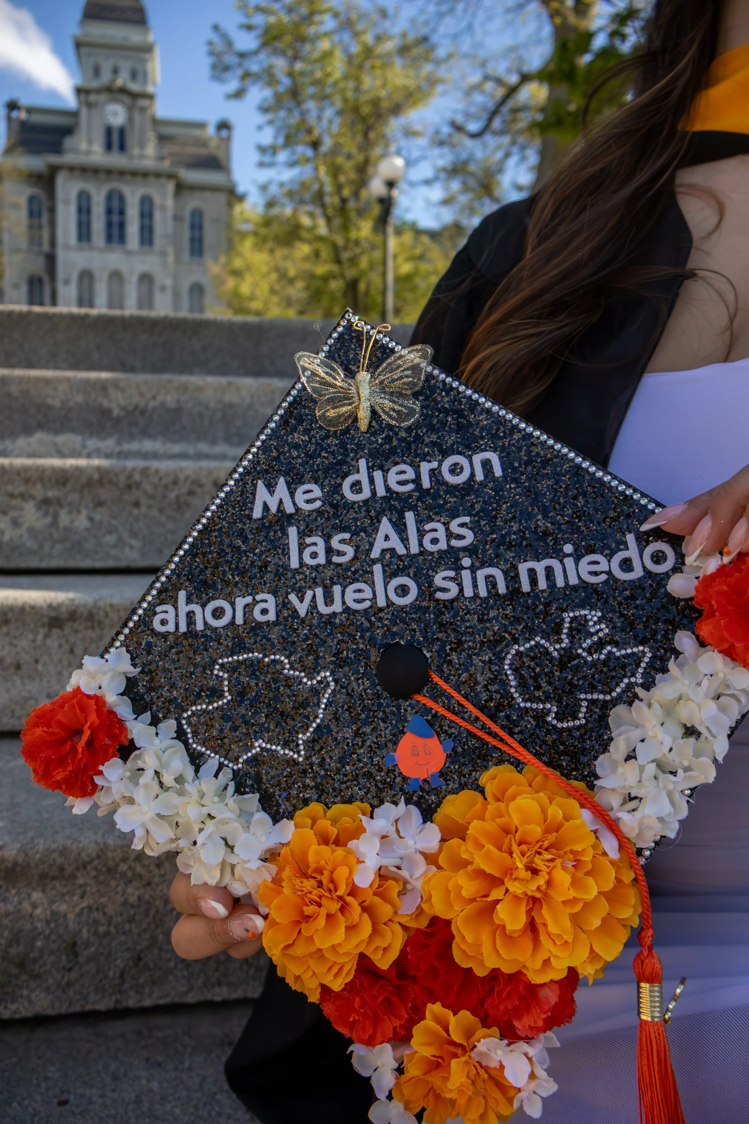 A graduation cap decorated with a golden butterfly, rhinestones, and colorful flower leis, held by a person wearing a black and white outfit with a yellow and black sash, with a historic building and trees in the background.