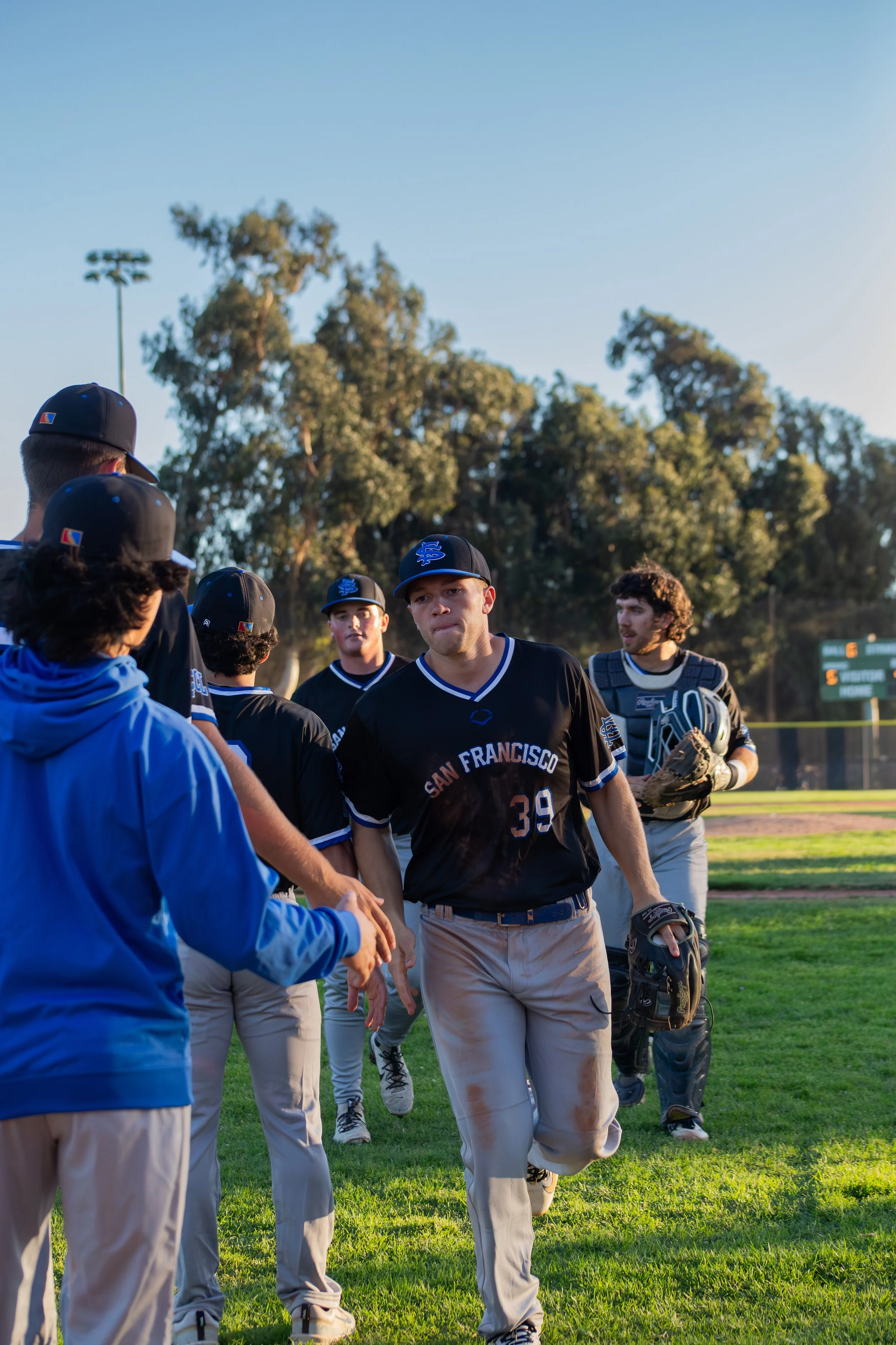 Baseball players from the San Francisco Giants team gather on the field, greeting, and shaking hands after a game, with some players holding gloves and wearing team uniforms and caps. The background features trees and a clear blue sky.