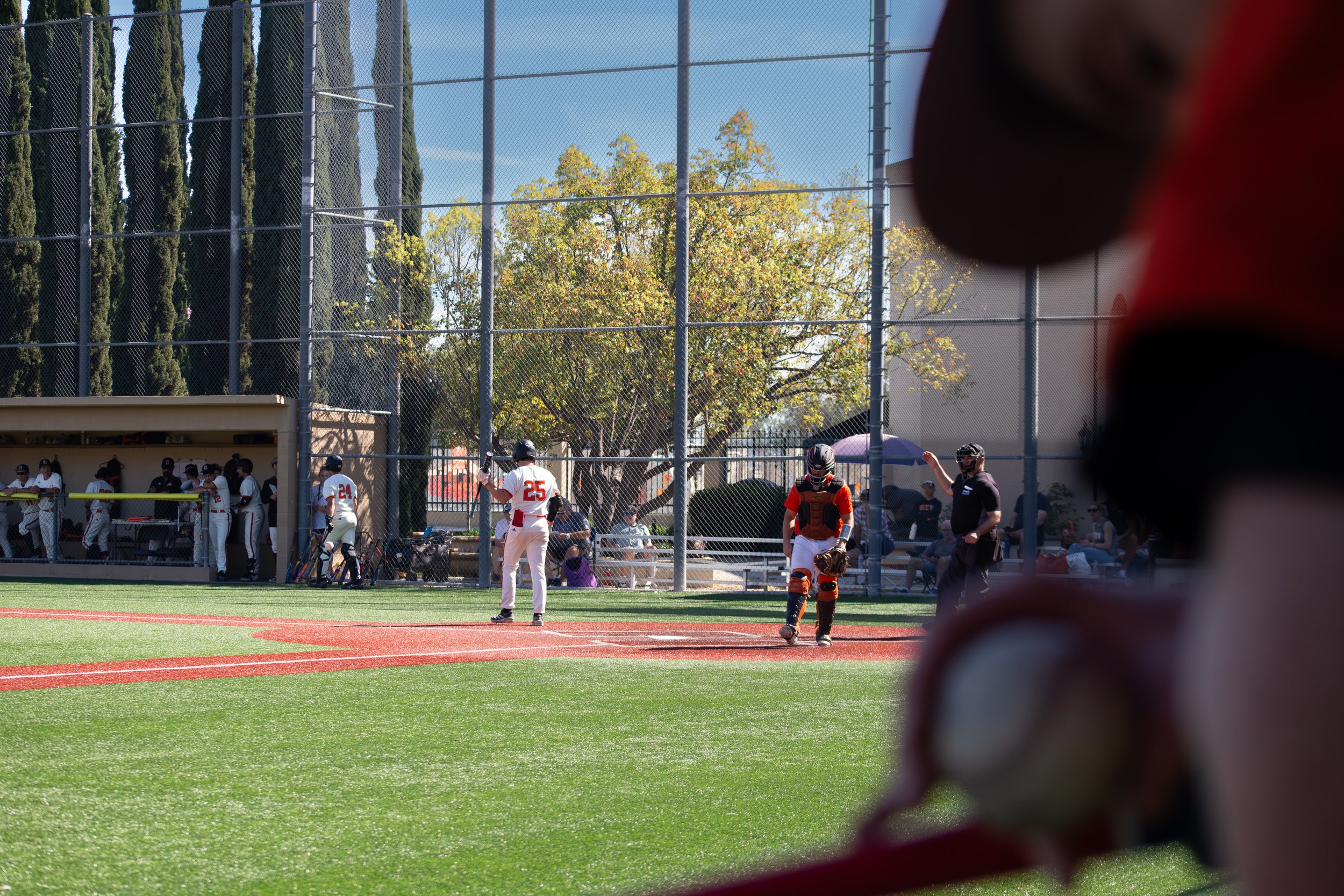 Baseball game in progress on a sunny day, with players in the dugout and on the field, viewed through a fence and a person's shoulder in the foreground.