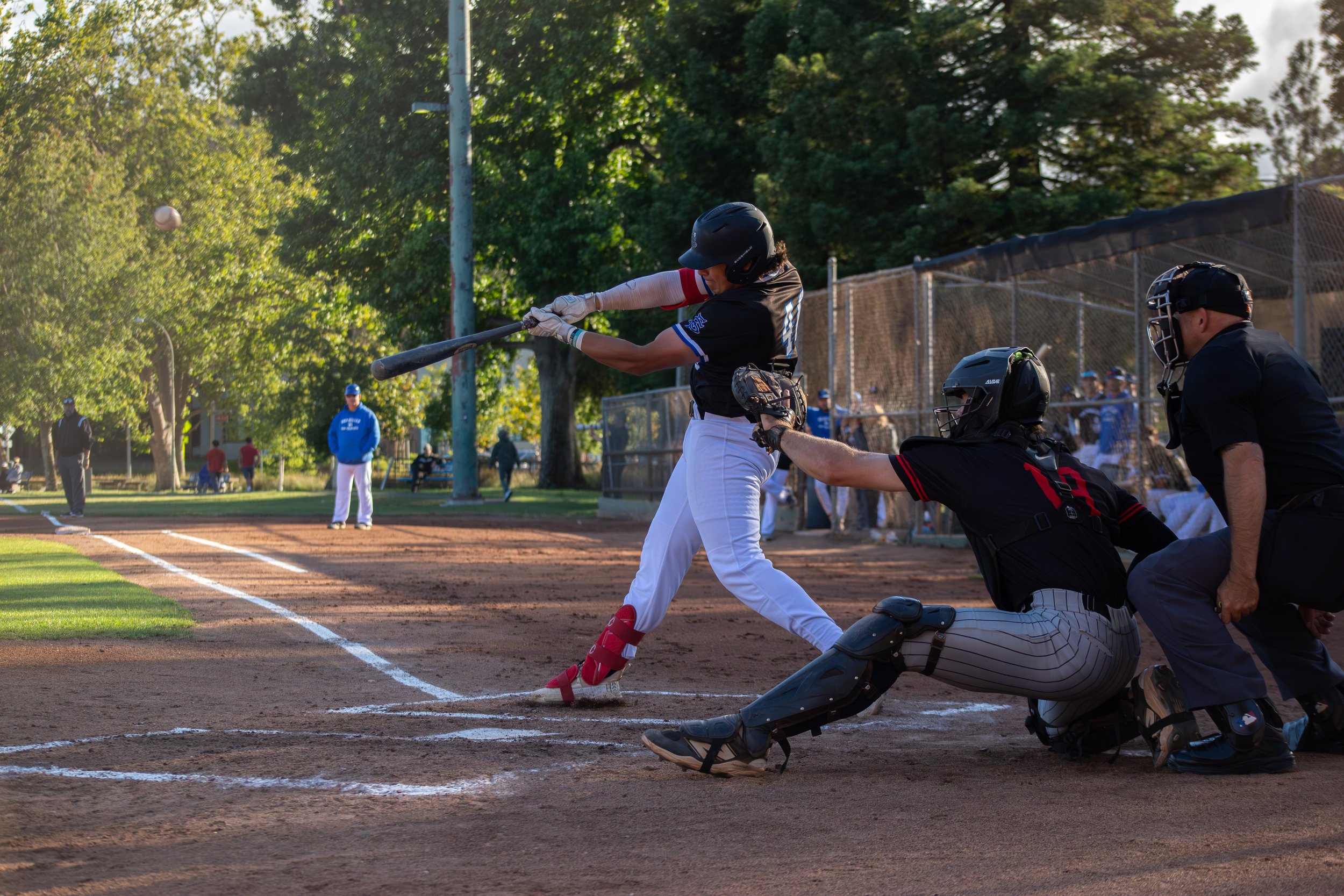 A baseball player swings a bat during a game at a park on a sunny day, with an umpire behind home plate and spectators watching in the background.