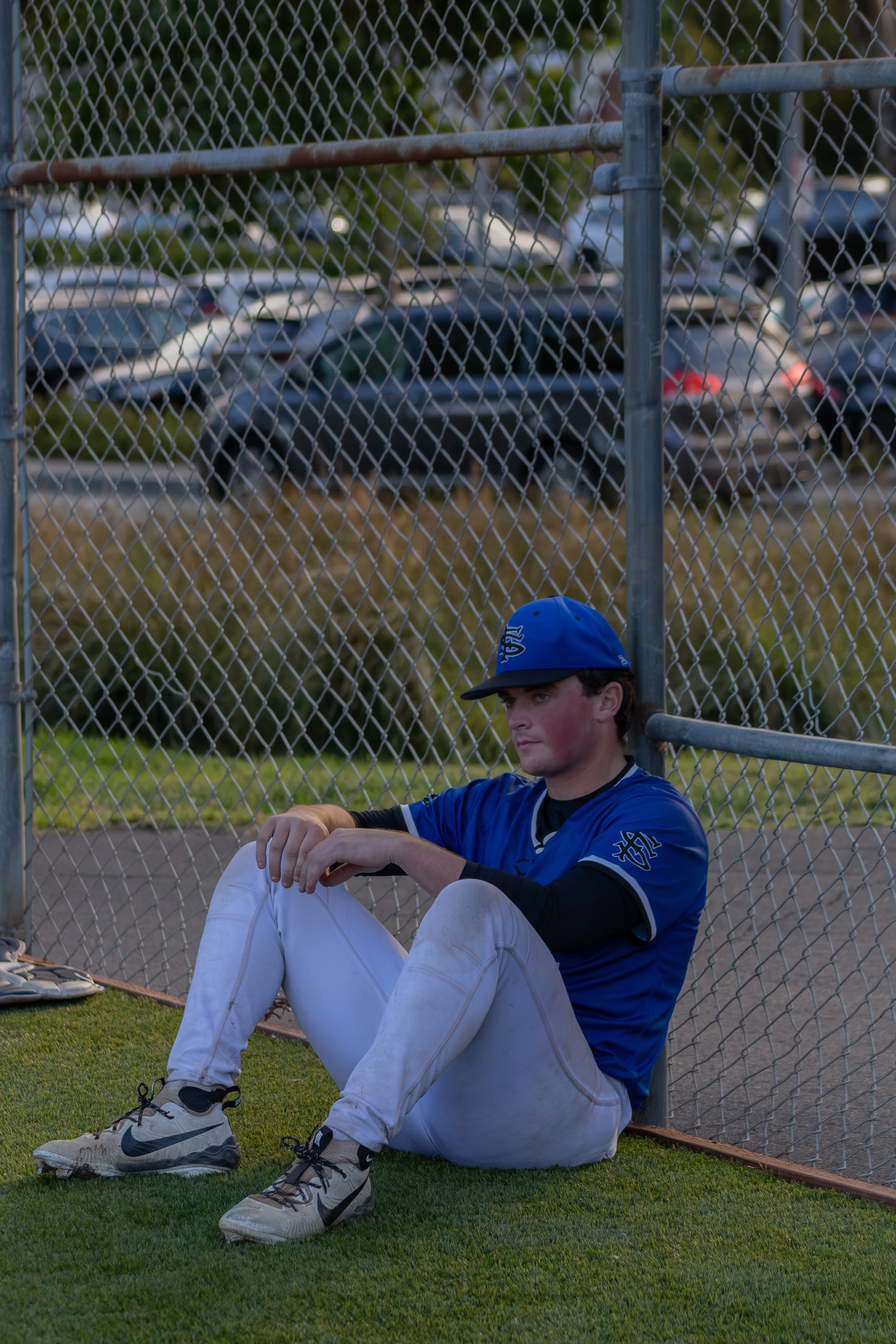 A young man sits on the grass at a baseball field, leaning against a chain-link fence, wearing a blue sports jersey, white pants, and a blue cap. The background shows a parking lot with several cars, slightly blurred.