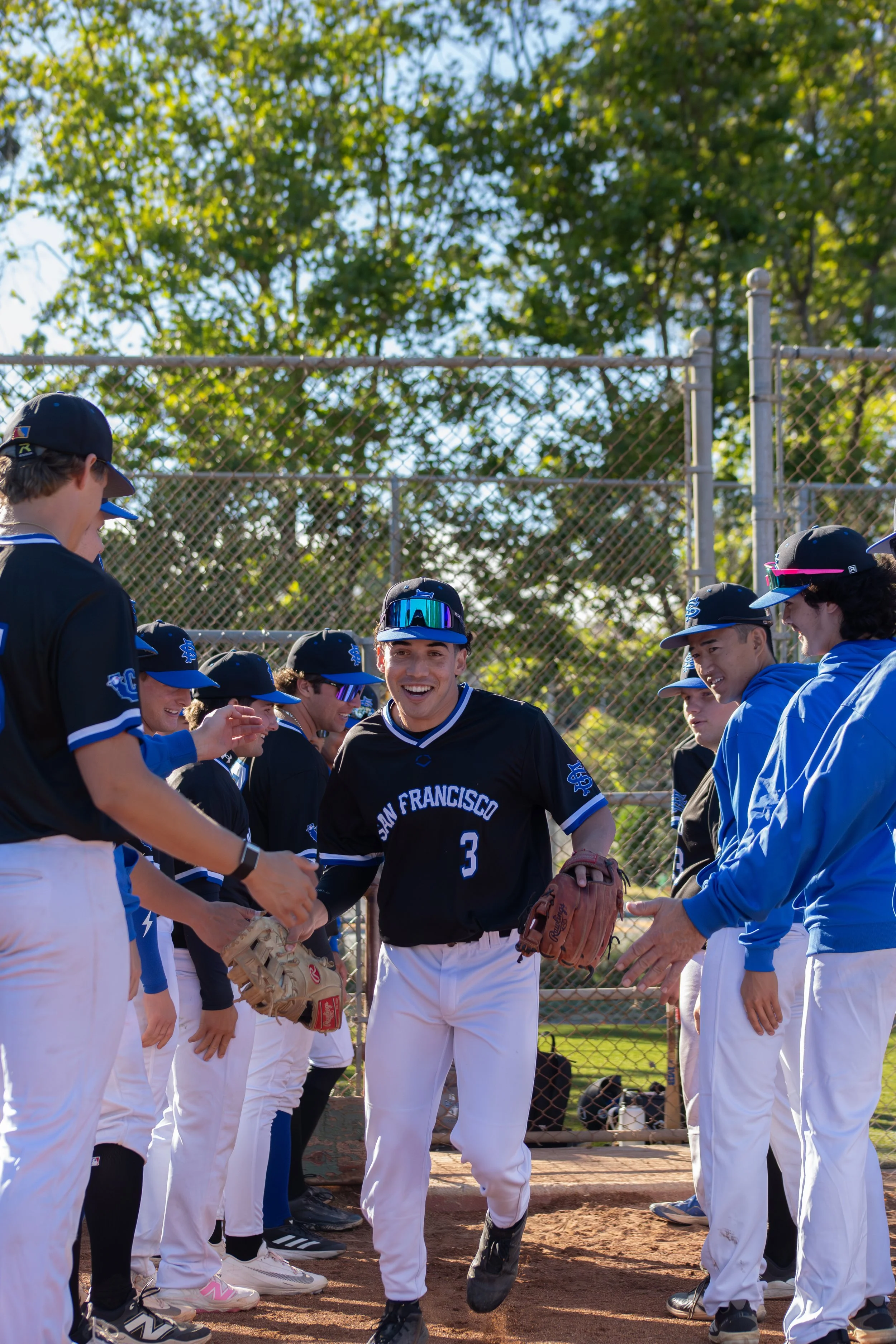 A group of young baseball players dressed in black and blue uniforms and white pants, gathered on a baseball field. They are smiling and appear to be celebrating or high-fiving each other. The background features a chain-link fence and tall green tre