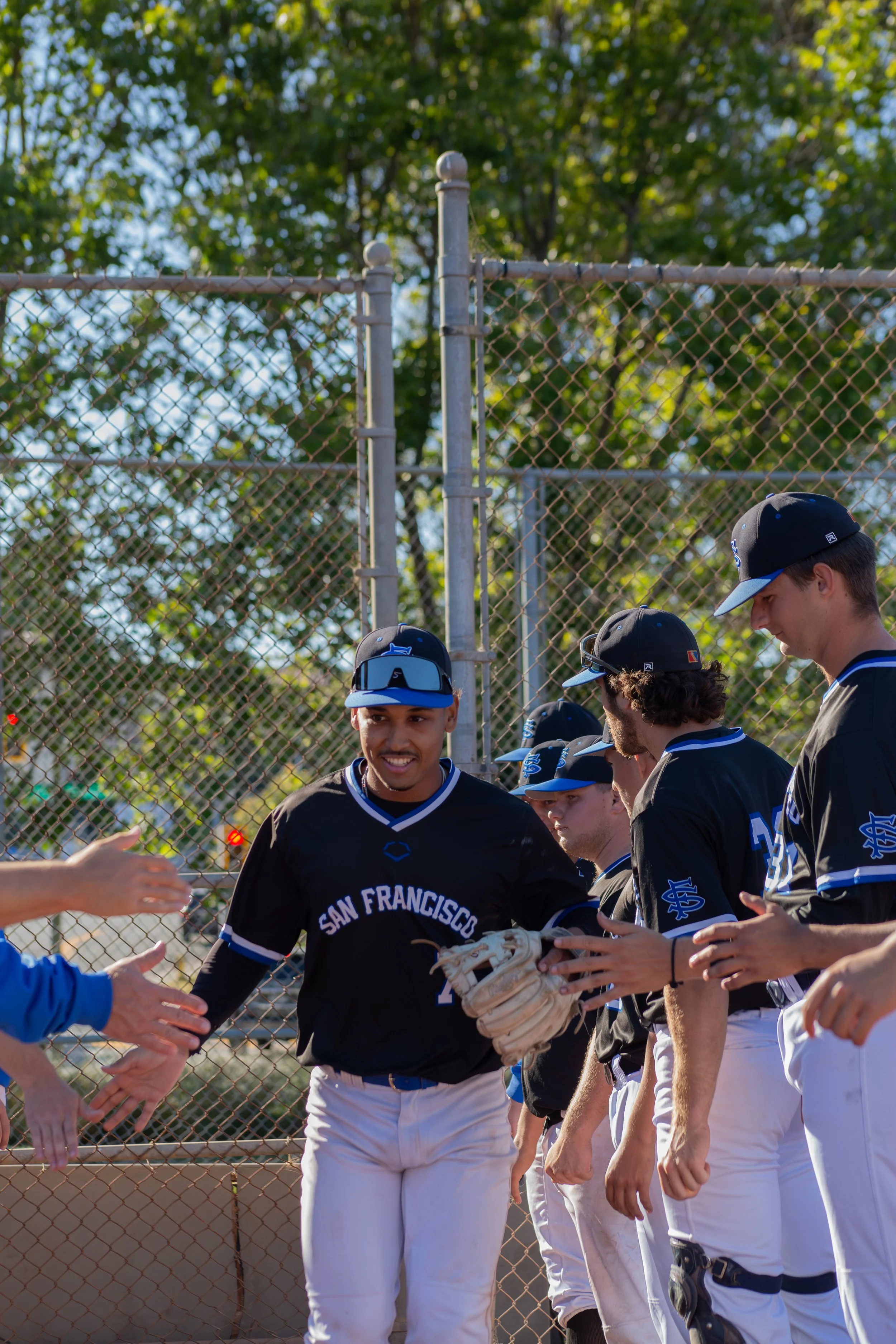 A group of baseball players in black uniforms with blue and white accents, wearing caps, are standing near a chain-link fence. One player, in the center, is smiling and holding a baseball glove, receiving high-fives from teammates and fans.