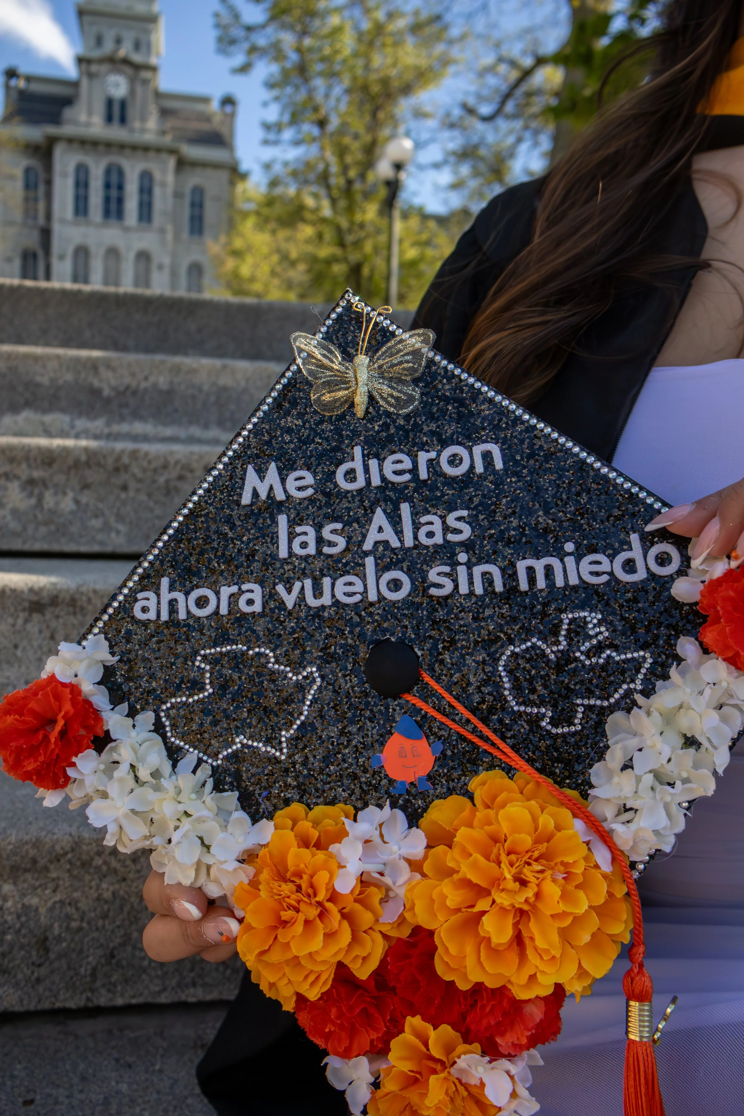 A graduation cap with a butterfly decoration, surrounded by colorful flowers, held by a person standing outdoors near a historic building with trees and a blue sky in the background.