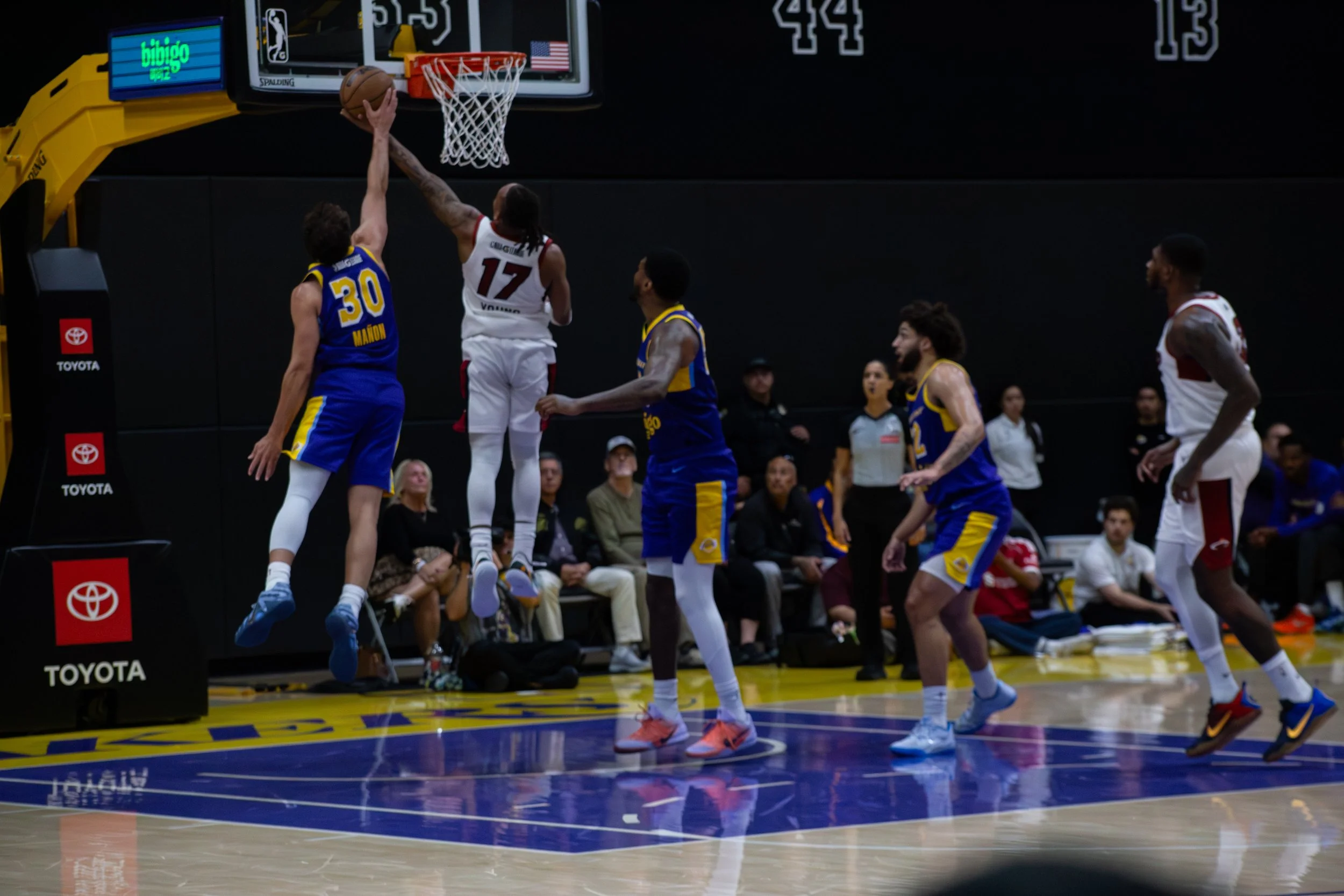 A basketball game showing a player in a white jersey with the number 17 attempting a shot near the hoop, while a player in a blue jersey with the number 30 defends. Several players are on the court, and spectators are seated in the background.