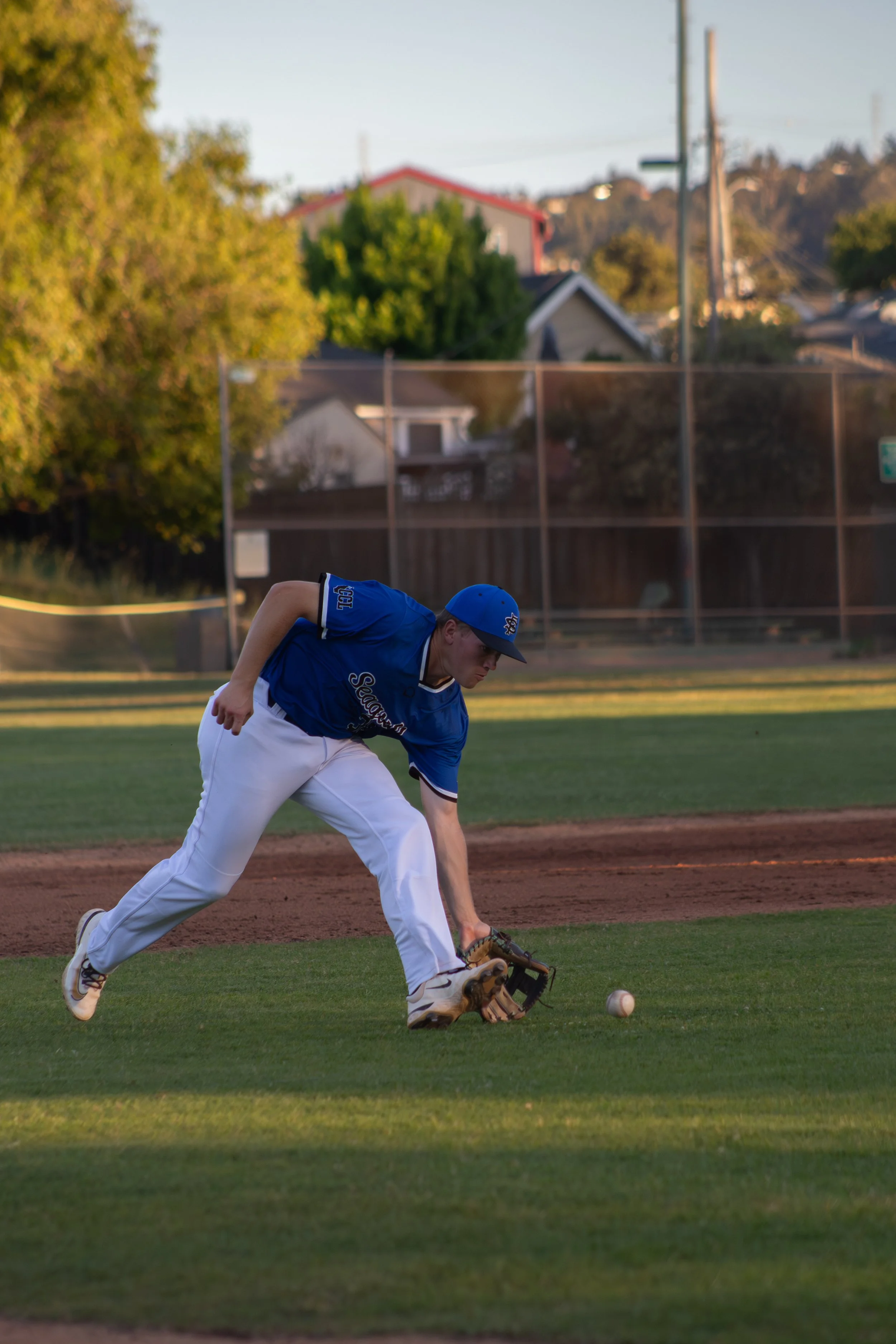 A baseball player in a blue jersey and white pants is fielding a ball on a baseball field, with grass, dirt, and a fence in the background, during late afternoon or early evening.