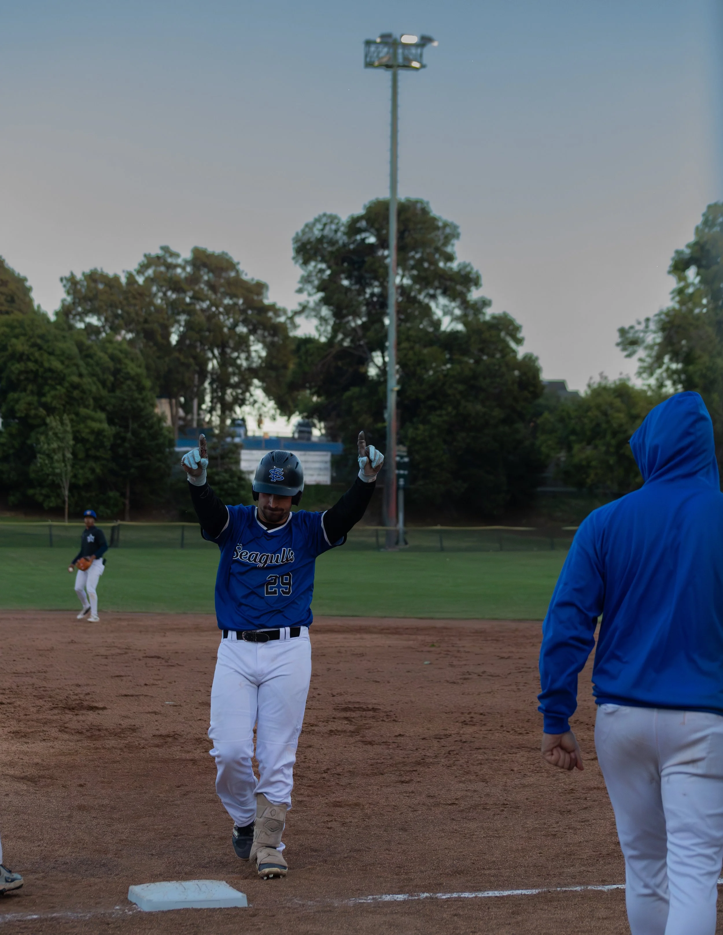 A baseball player in a blue jersey with the number 29 is walking on a baseball field with his arms raised, while another person in a blue hoodie stands nearby. In the background, a player in white pants and a dark jersey is standing on the field. Tre