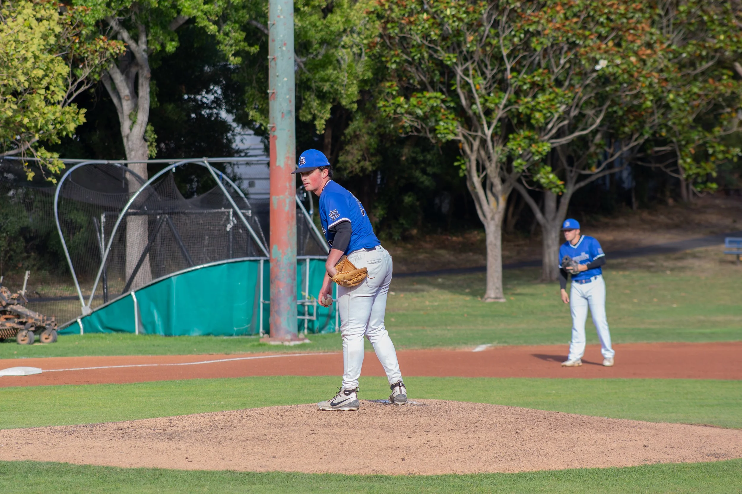 Two baseball players practicing on the field, one pitching from the mound and another in the outfield, near trees and a fence.