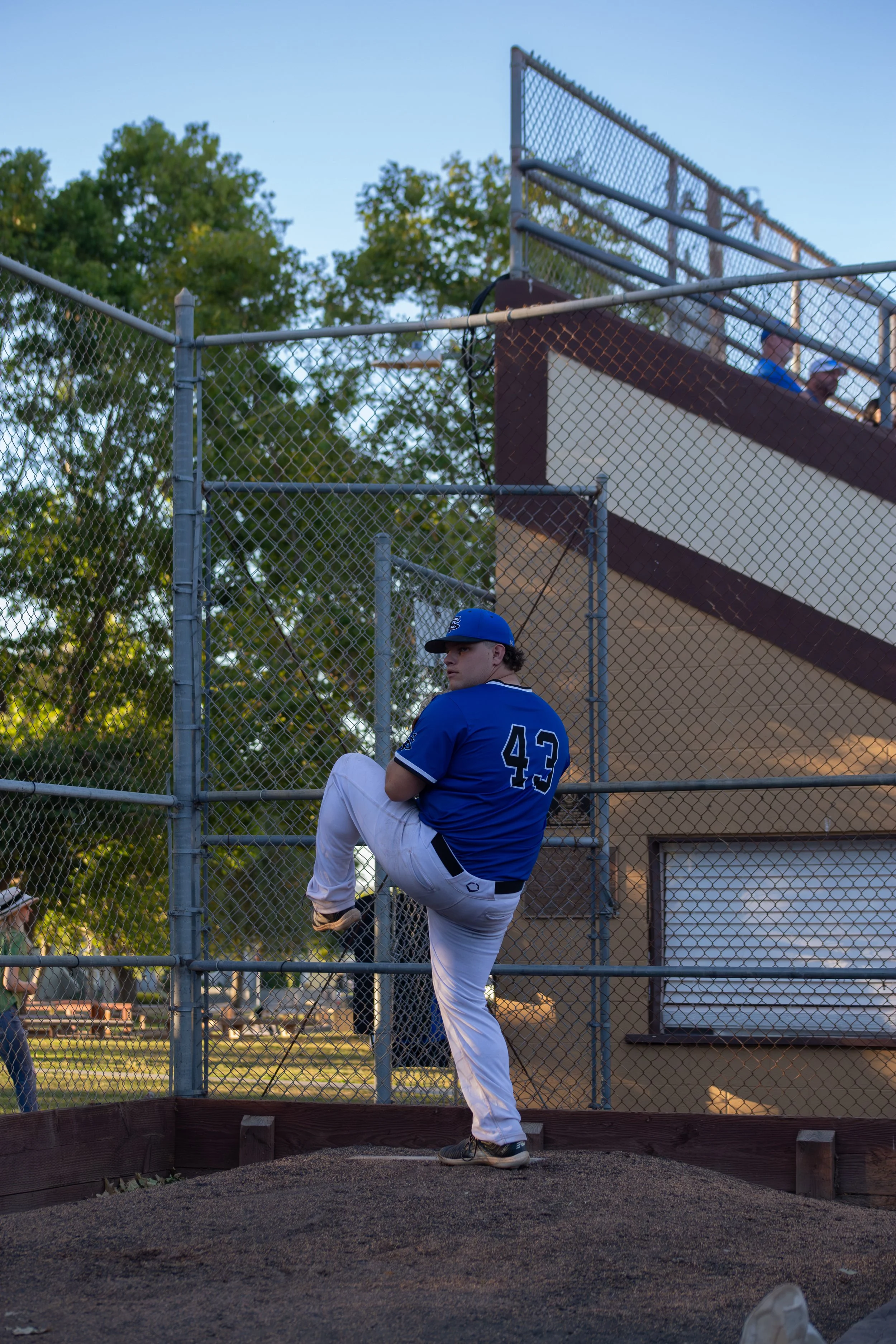 A baseball player in a blue jersey with the number 43 is warming up on a bullpen mound, with one leg lifted, behind a chain-link fence at a baseball field during the daytime.