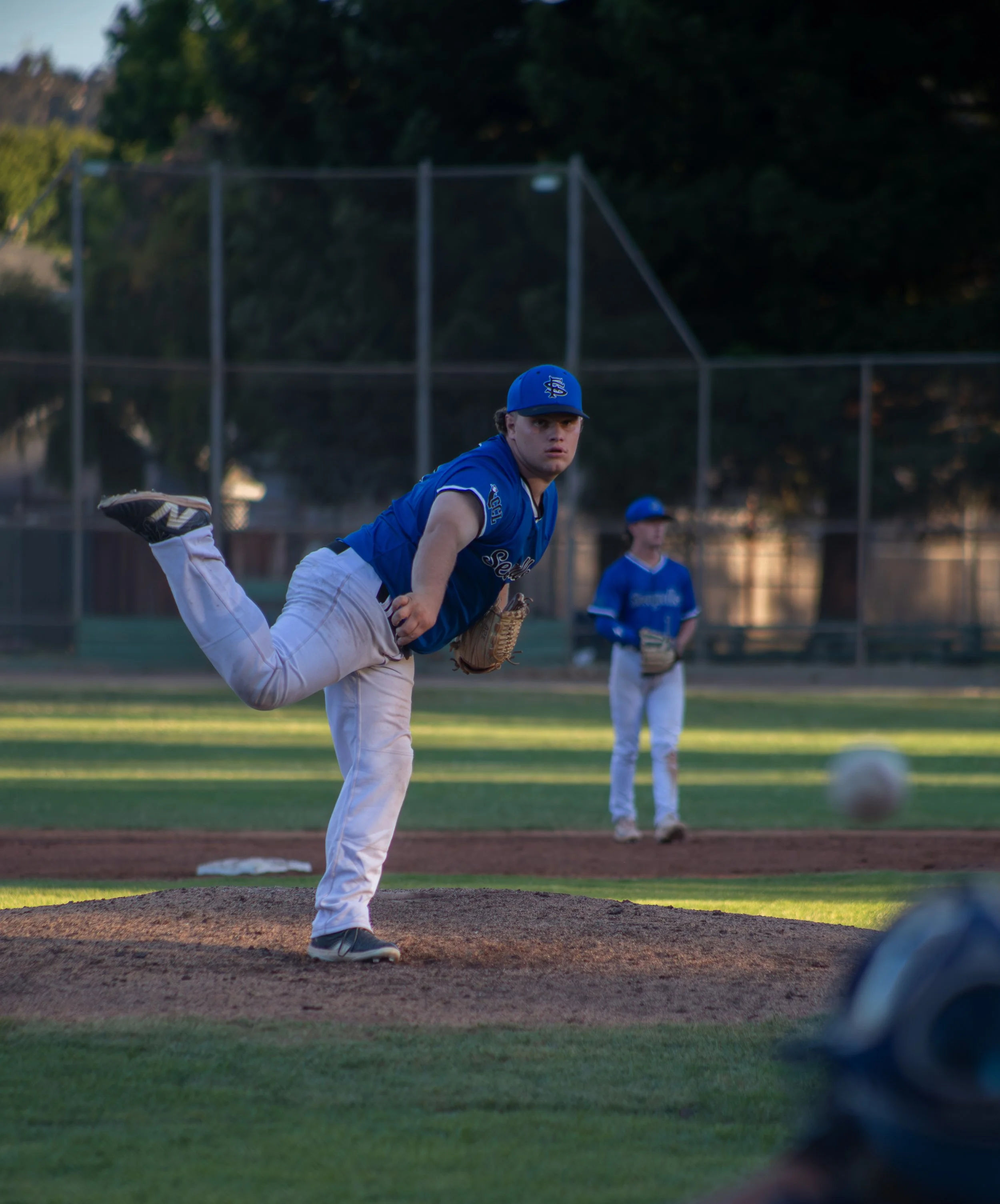 A young baseball player in a blue uniform and cap is pitching on the mound during a game. Other players and a chain-link fence are visible in the background, with trees and a sky at sunset.
