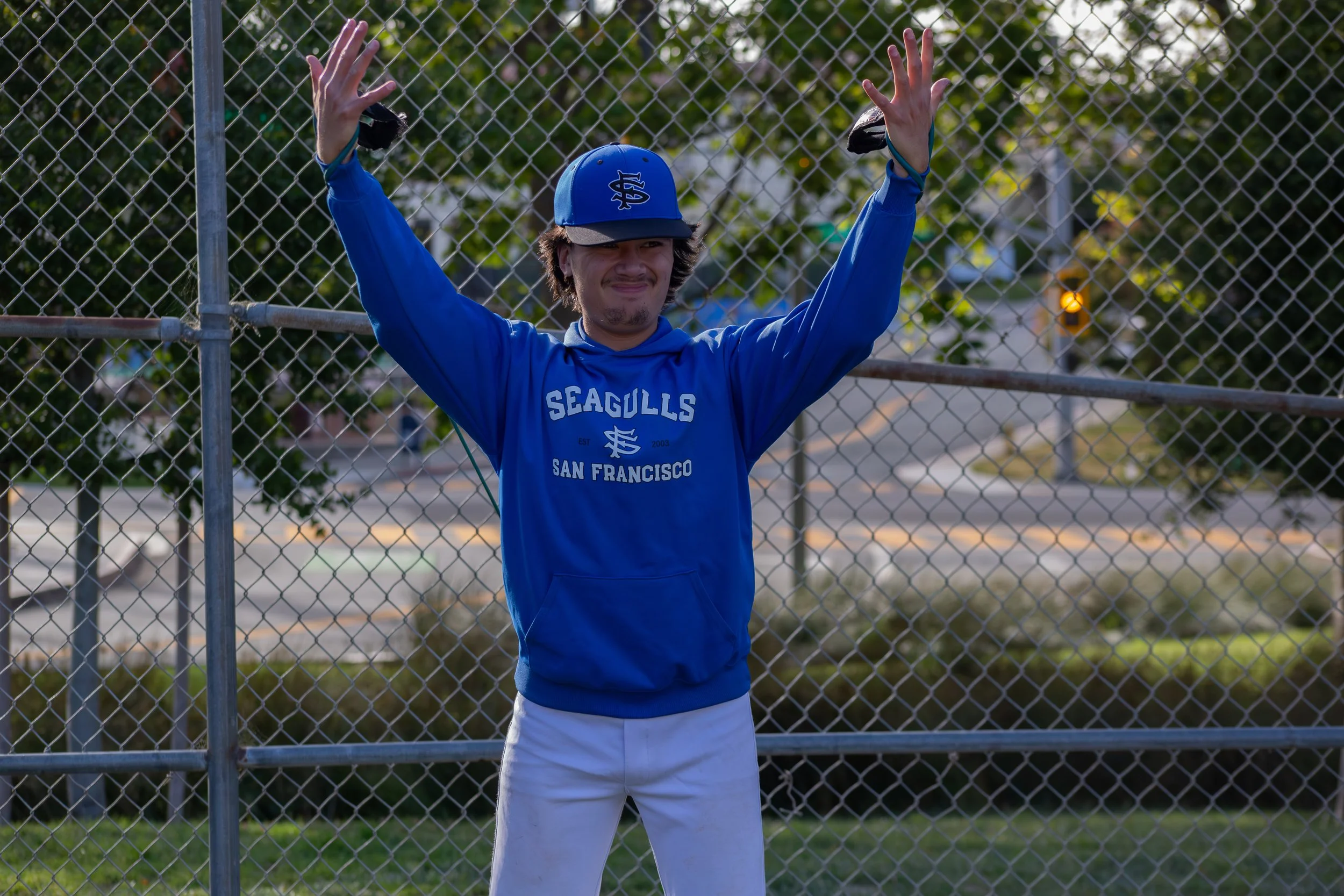 A young man in a blue hoodie with 'Seagulls San Francisco' printed on it and a blue cap stands behind a chain-link fence with his hands raised and enjoying the outdoor setting.