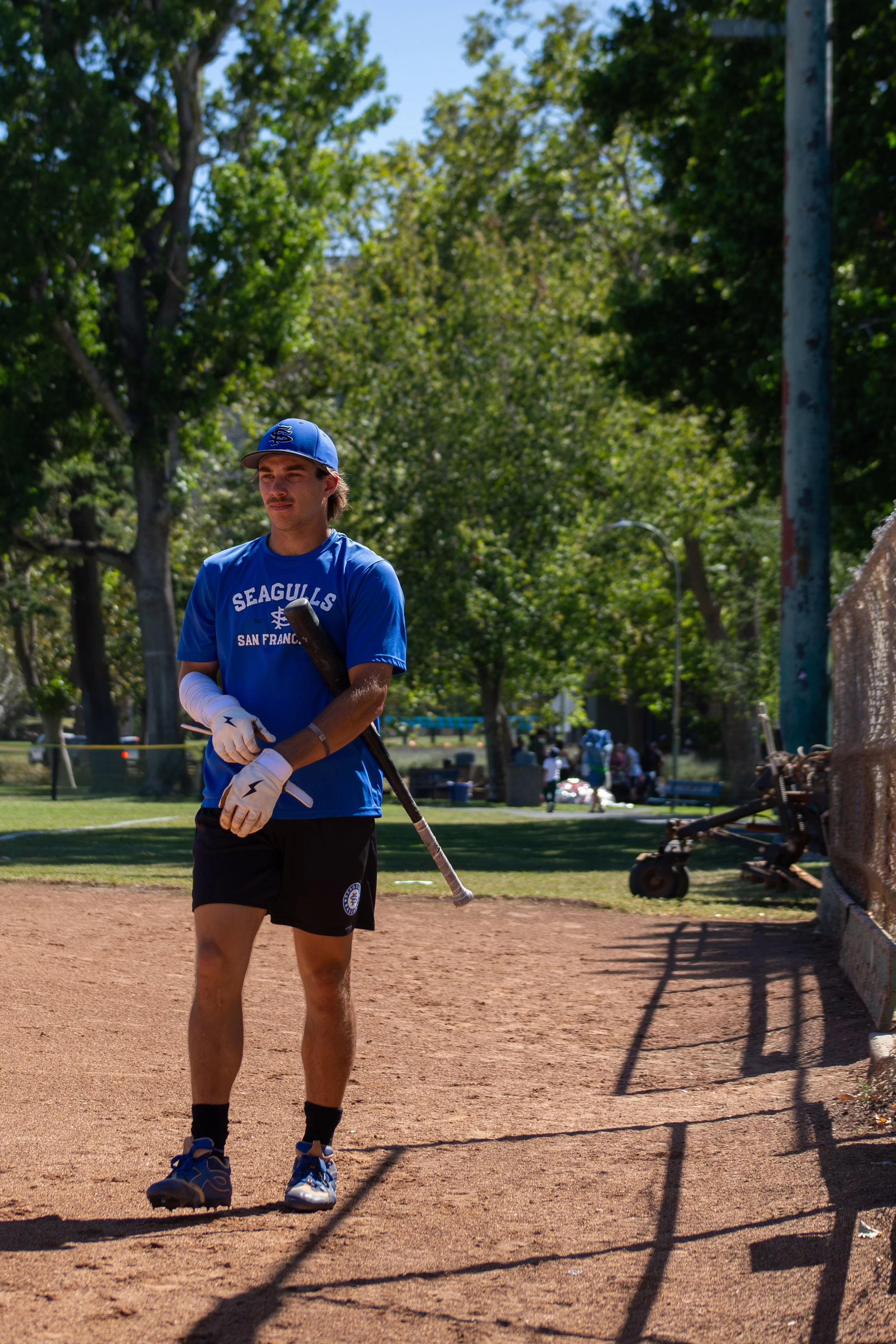 A young man in a blue San Francisco Giants t-shirt, black shorts, and blue sneakers stands on a baseball field holding a baseball bat. He is wearing a blue cap and white gloves. The background shows trees, a park, and a few people in the distance.