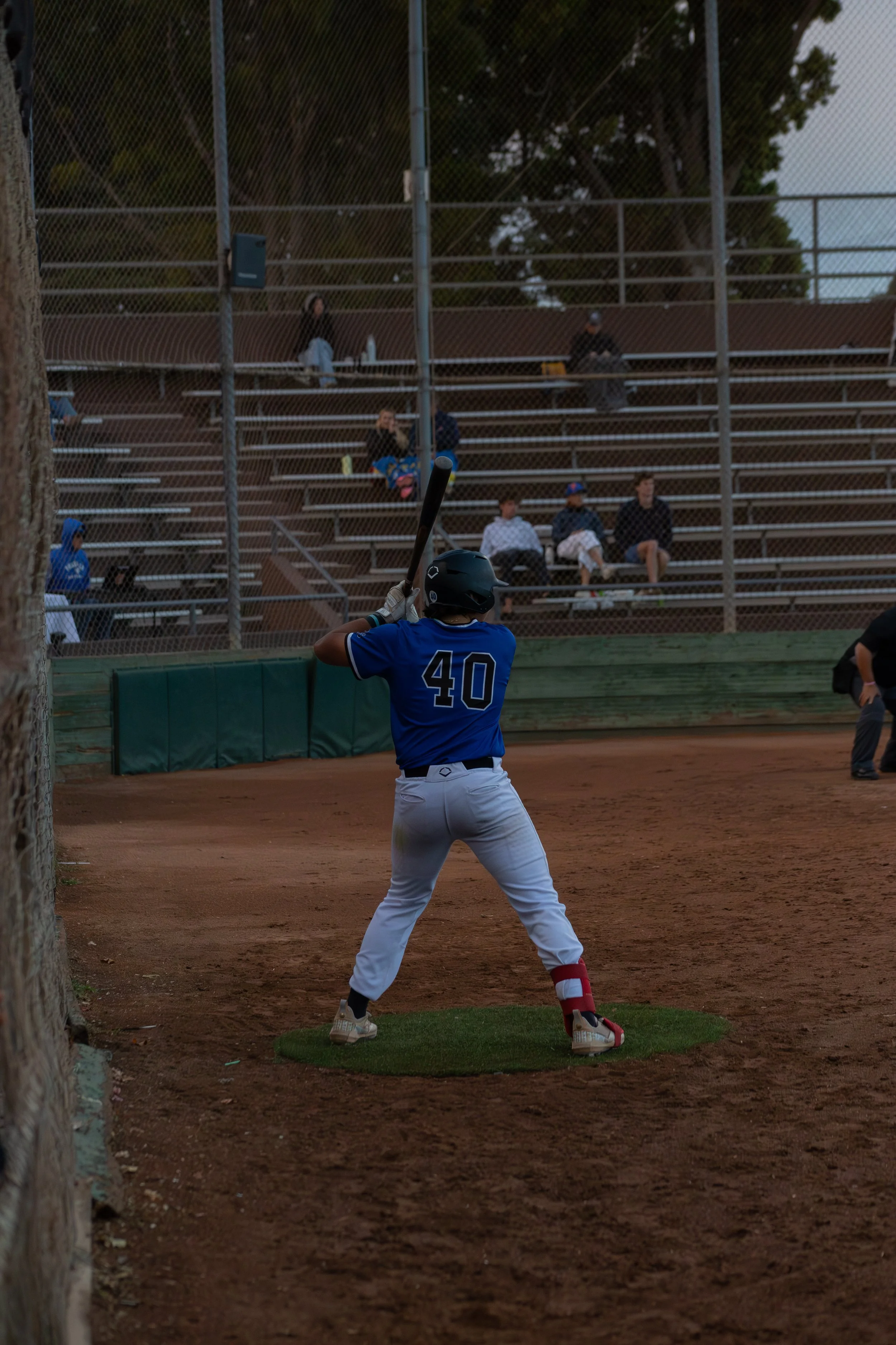 A baseball player in a blue jersey with the number 40 is positioned at bat on a baseball field, preparing to swing. There are spectators sitting on bleachers behind a chain-link fence, with some watching the game. The scene appears to be during the e