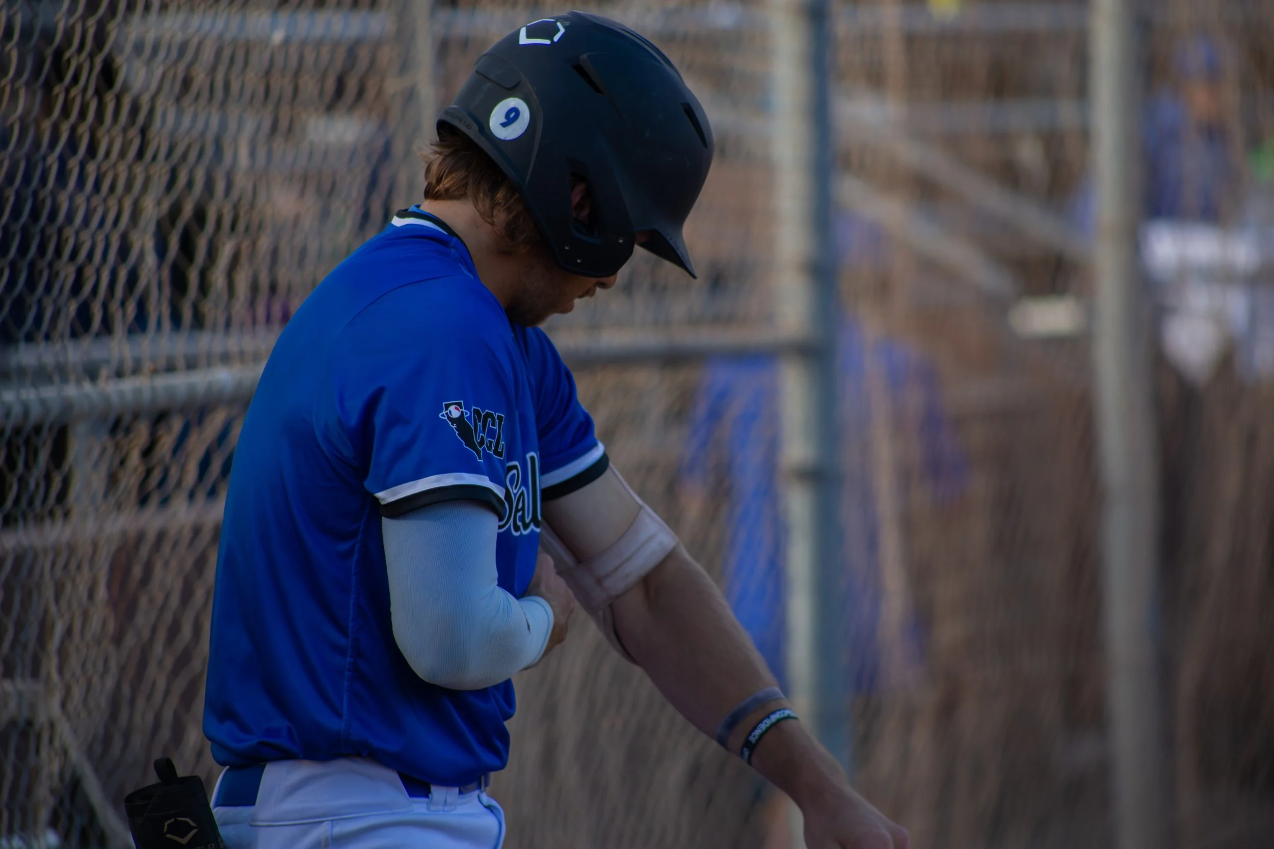 A baseball player in a blue uniform and black helmet standing near a chain-link fence with a wooden bat.