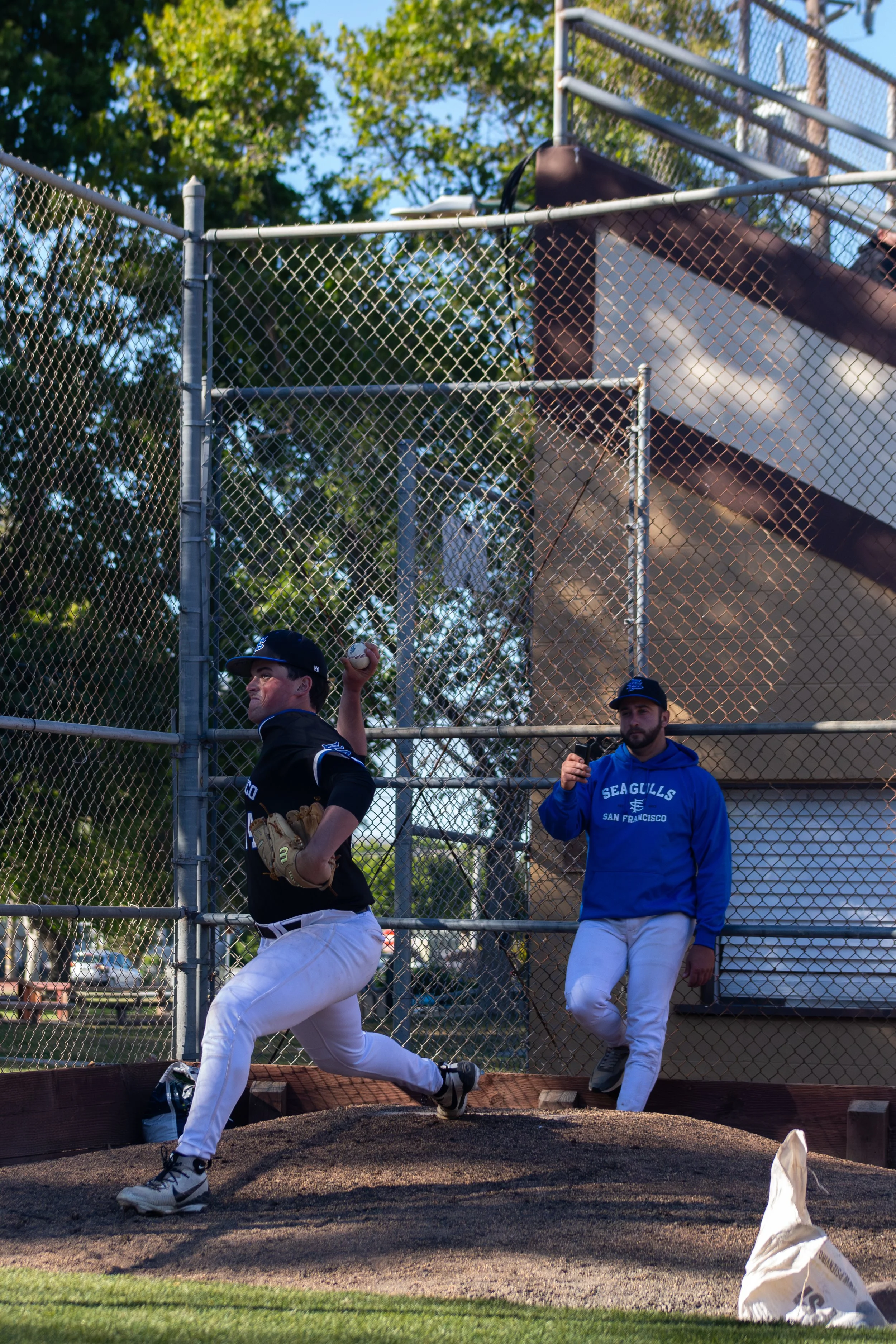 Two baseball players practicing pitching on a baseball field with a chain-link fence, trees, and a slide in the background; one player is in mid-throw and the other is holding a phone, both wearing athletic gear.