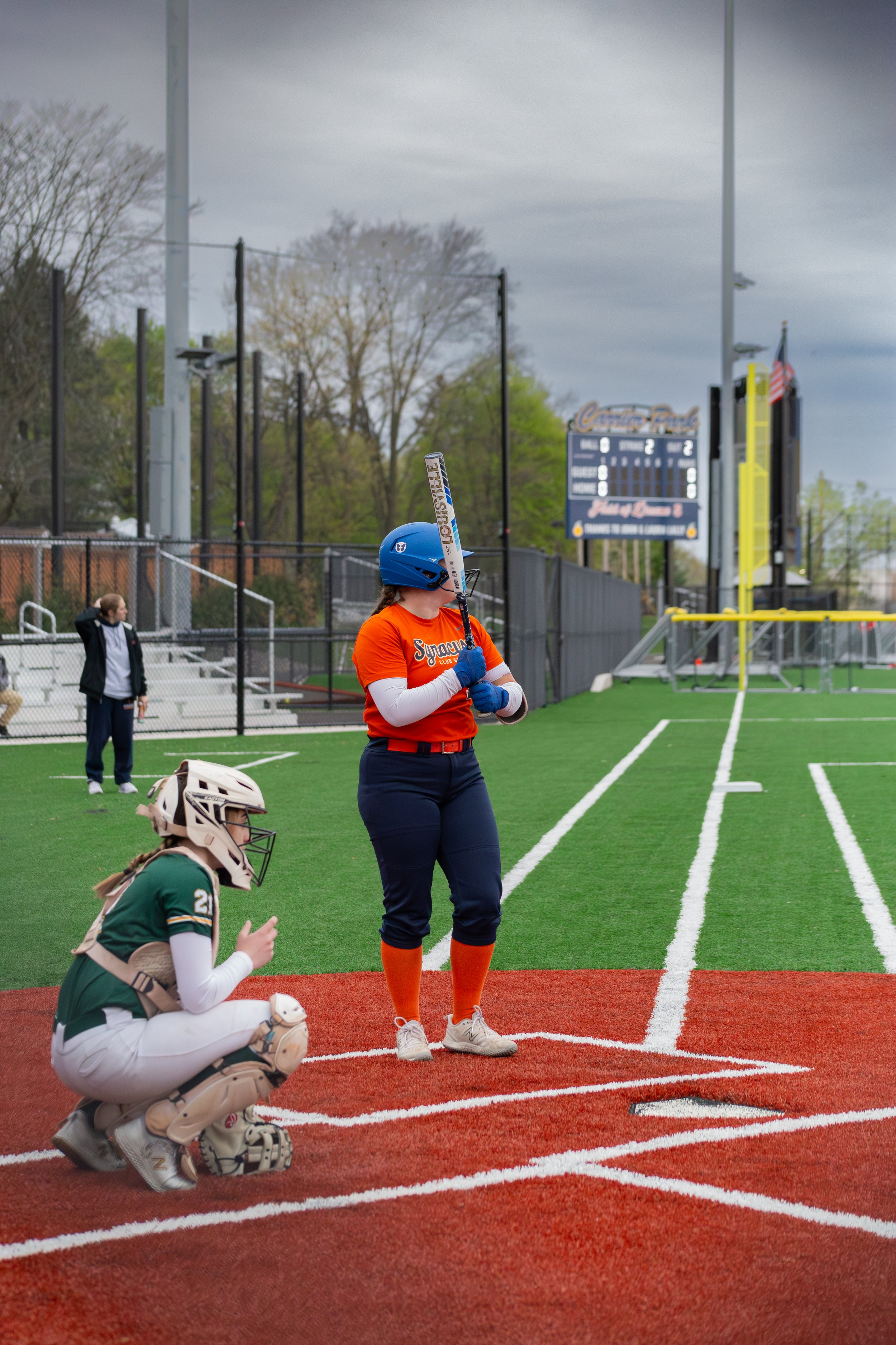 A female softball batter in an orange uniform and blue helmet holding a bat at home plate during a game on a baseball field, with a catcher in green and white kneeling in front and a scoreboard in the background.