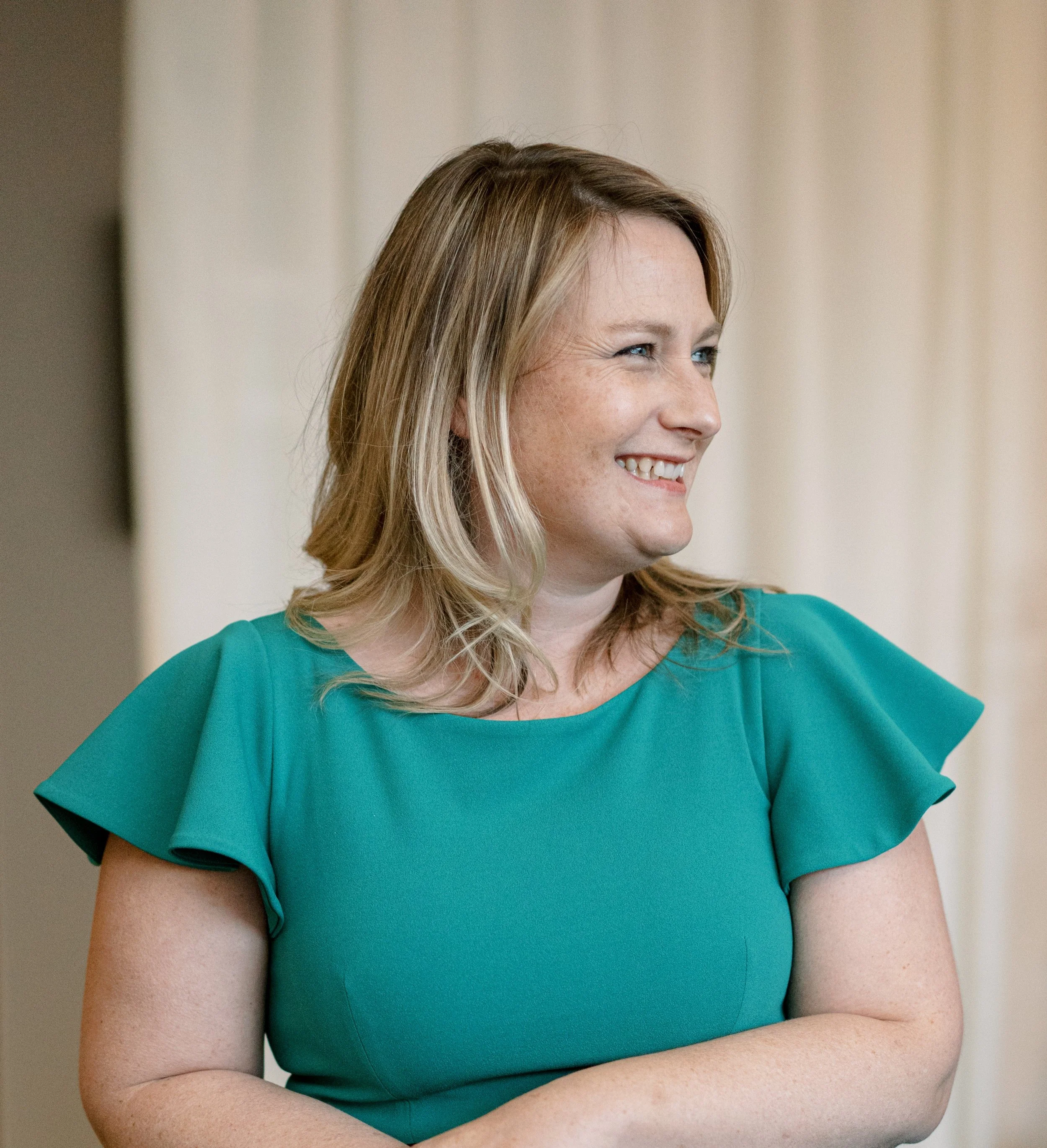 Close-up of a woman smiling, wearing a teal dress with ruffle details on the sleeves, standing indoors with a neutral background.