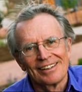 Close-up of a smiling older man with gray hair and glasses, wearing a blue shirt, outdoors with blurred trees in the background.